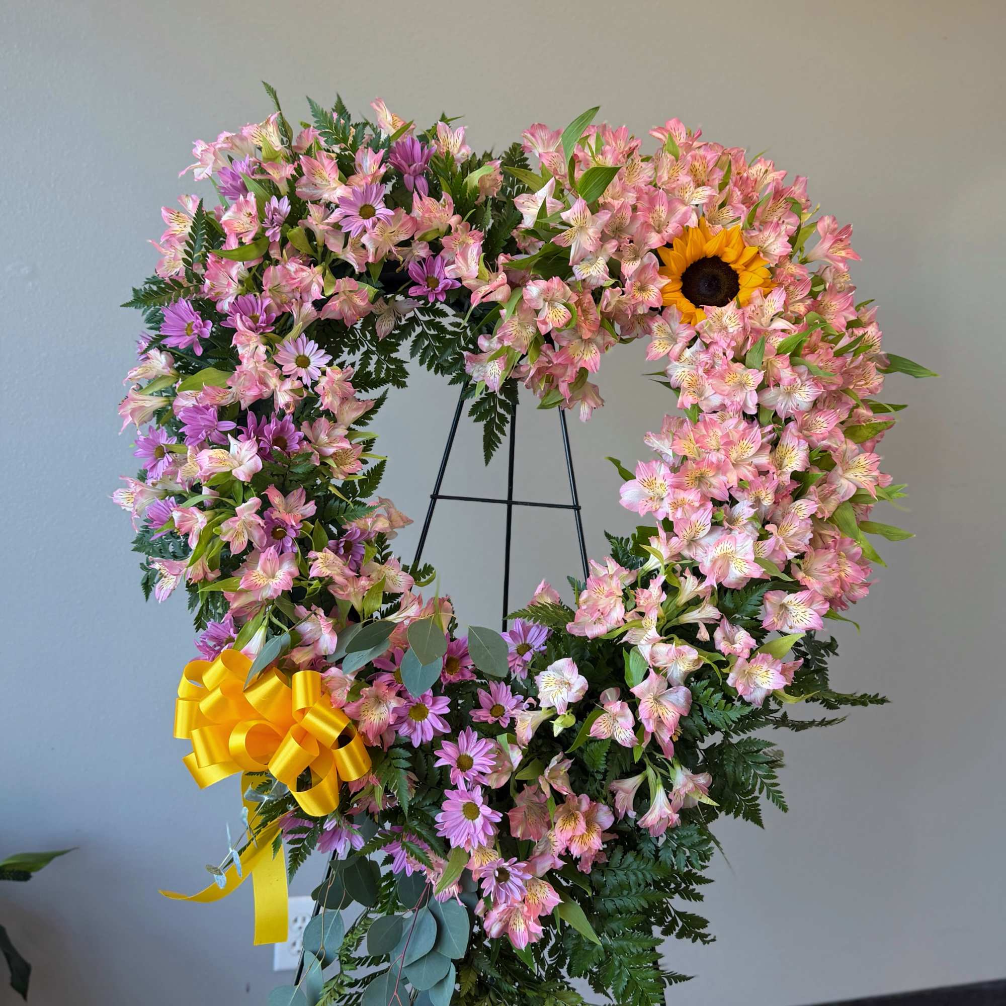 Heart-shaped standing wreath of pink flowers with a yellow sunflower and bow on a metal easel