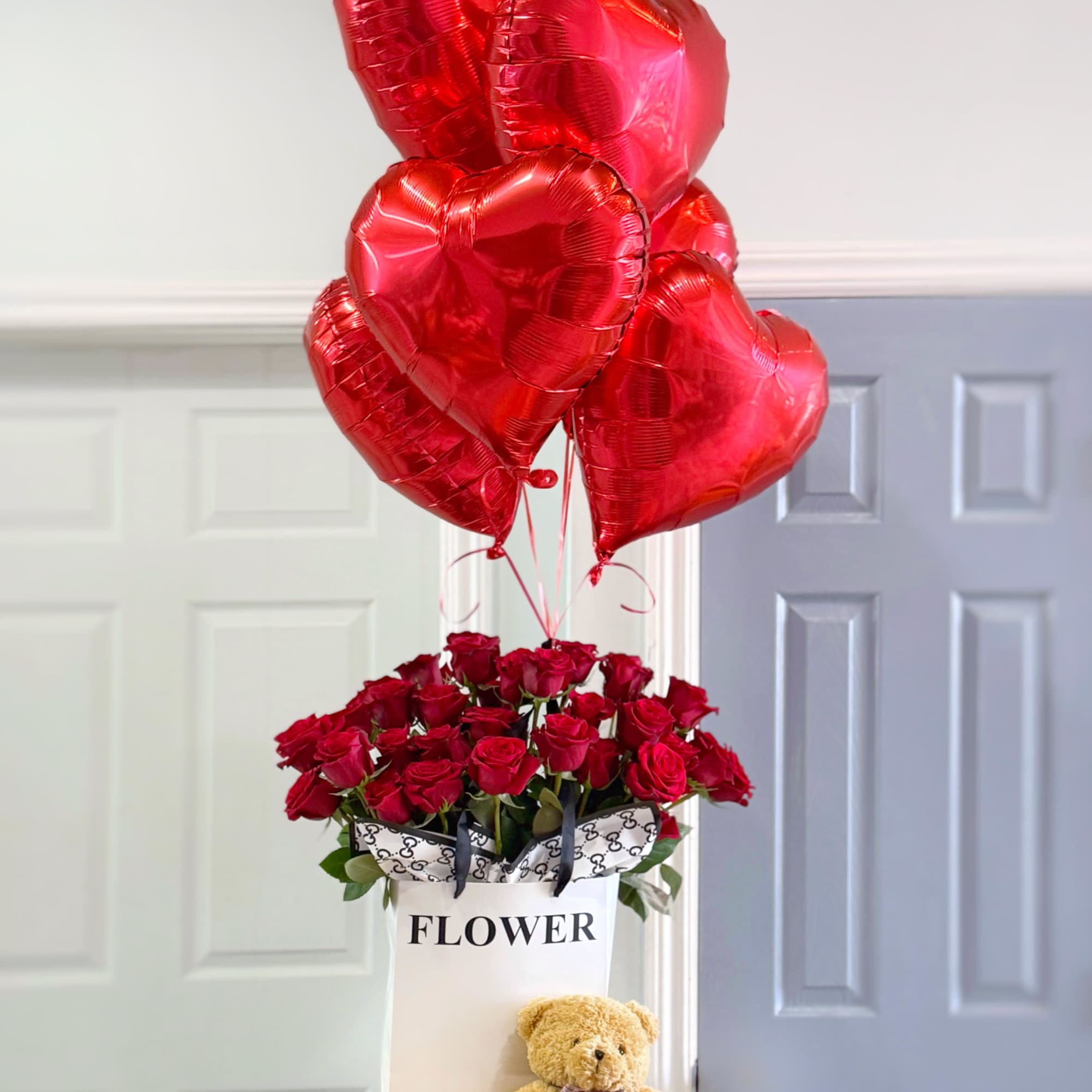 Red roses in a white gift bag with heart balloons and a teddy bear