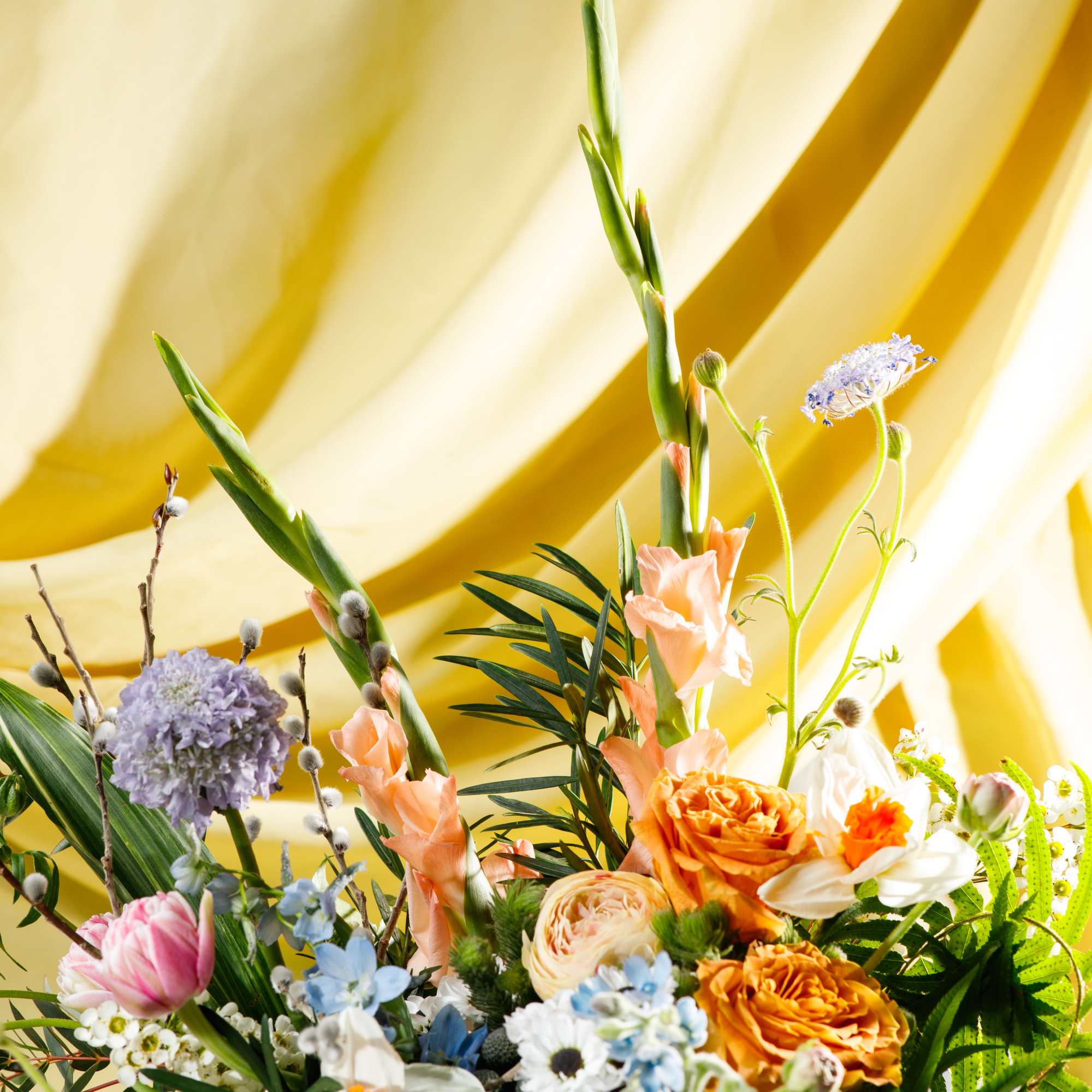 Colorful mixed bouquet with peach roses, tulips, and assorted blooms against a yellow fabric backdrop