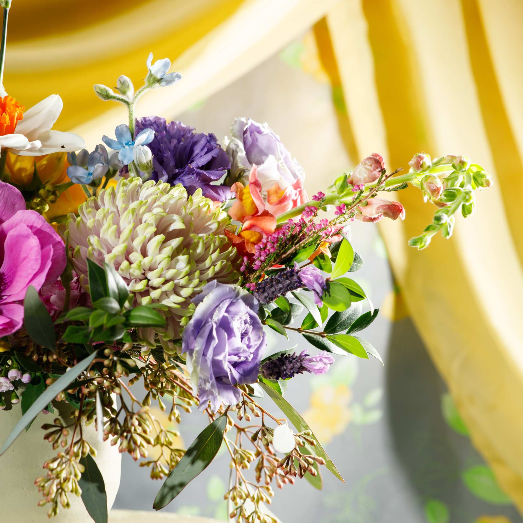 Mixed bouquet with purple lisianthus, chrysanthemum, and pink flowers in a white vase on a draped table.