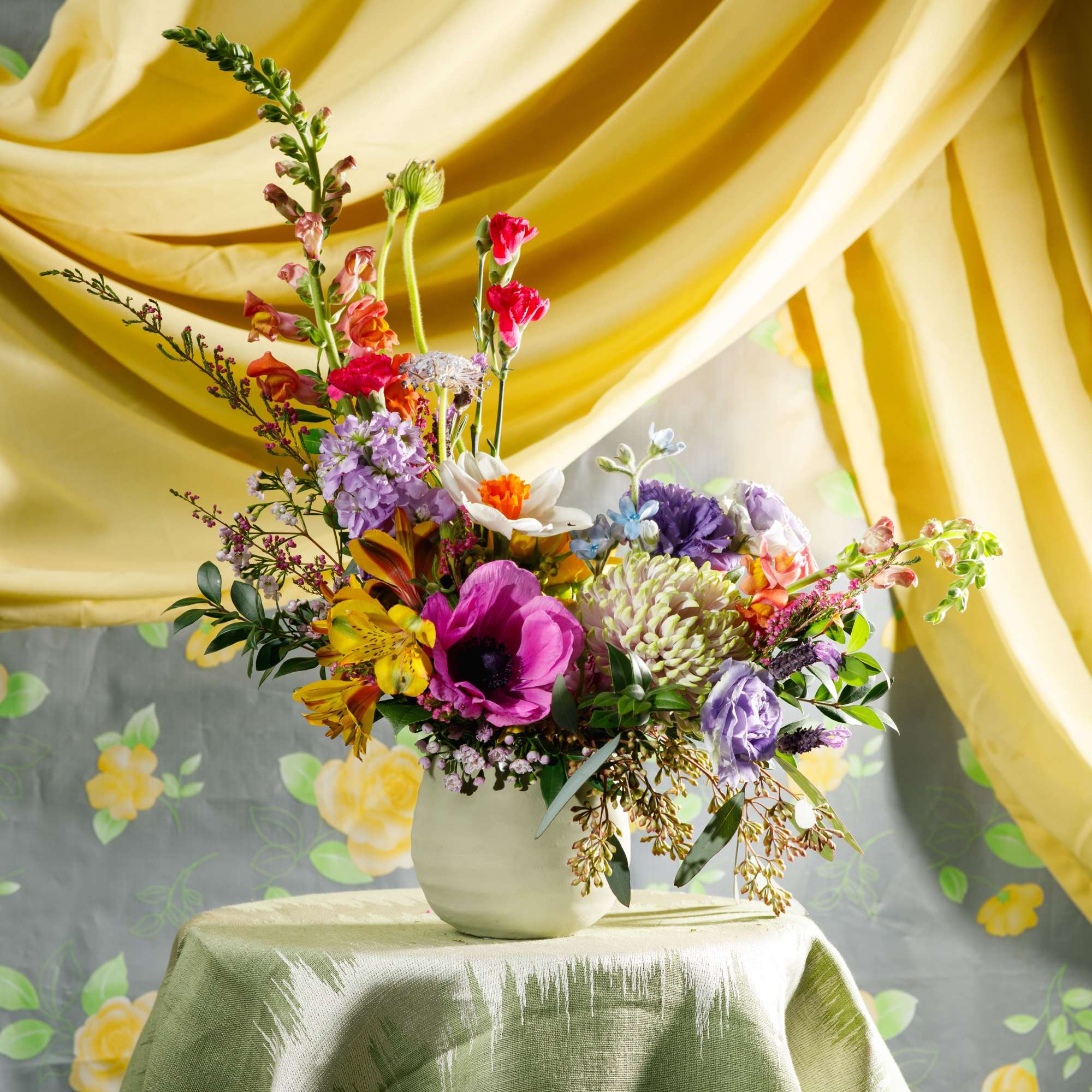 Colorful mixed flower arrangement in a white vase on a draped table against yellow fabric.