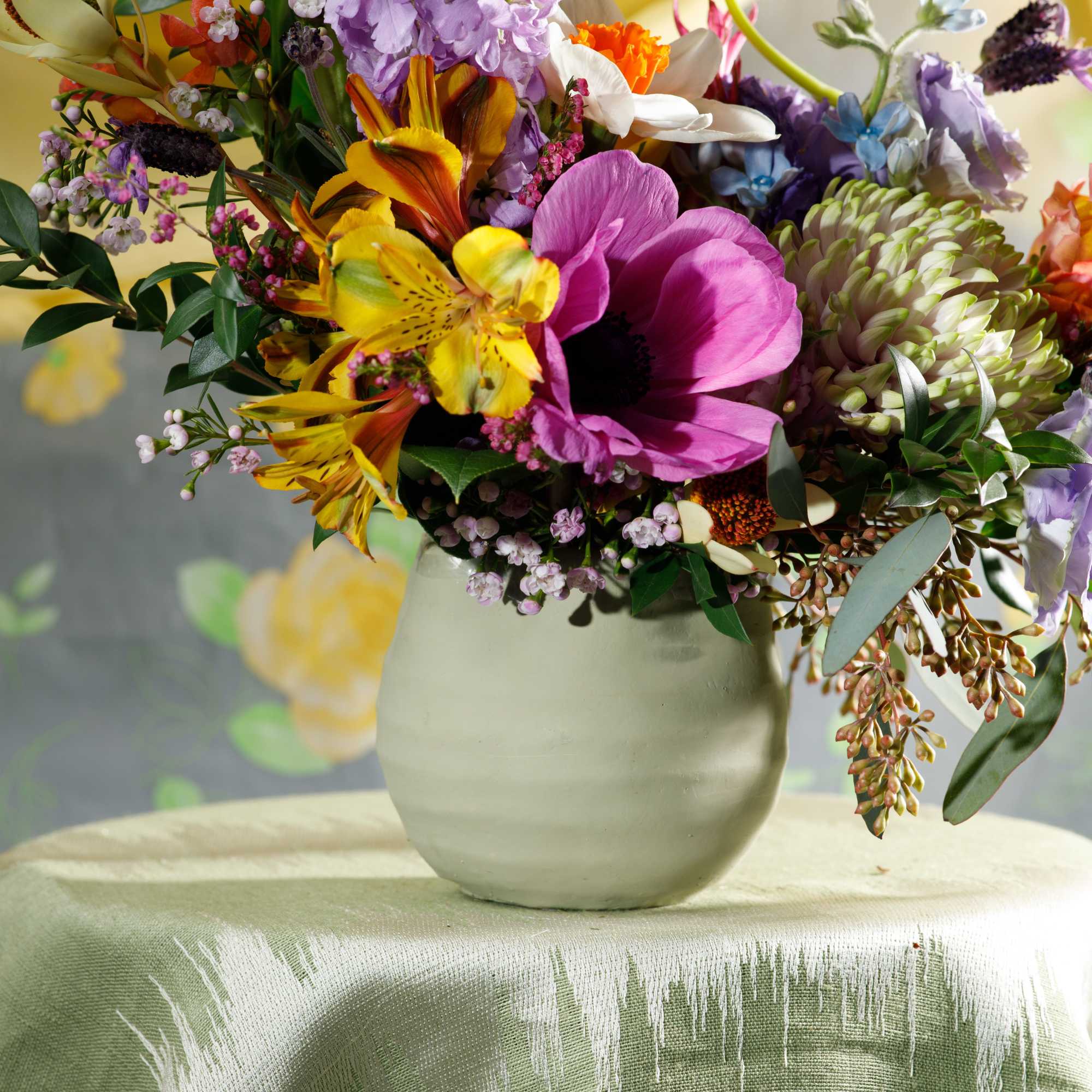 Bright mixed bouquet of purple, yellow, and orange flowers in a pale ceramic vase on a draped table.