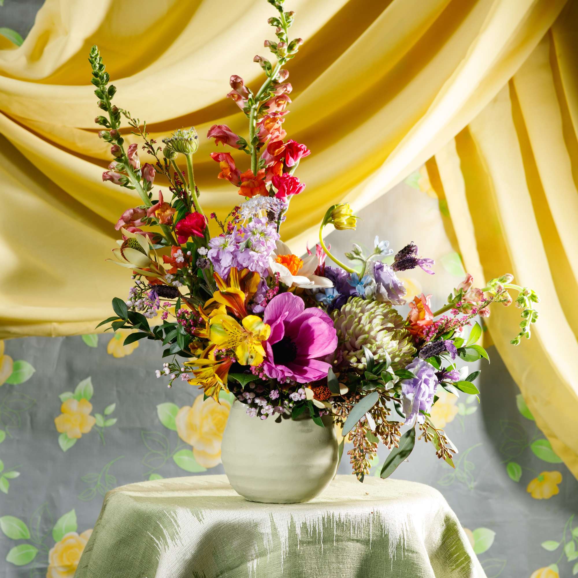 Tall mixed-color flower arrangement in a pale ceramic vase on a draped table with yellow fabric backdrop.