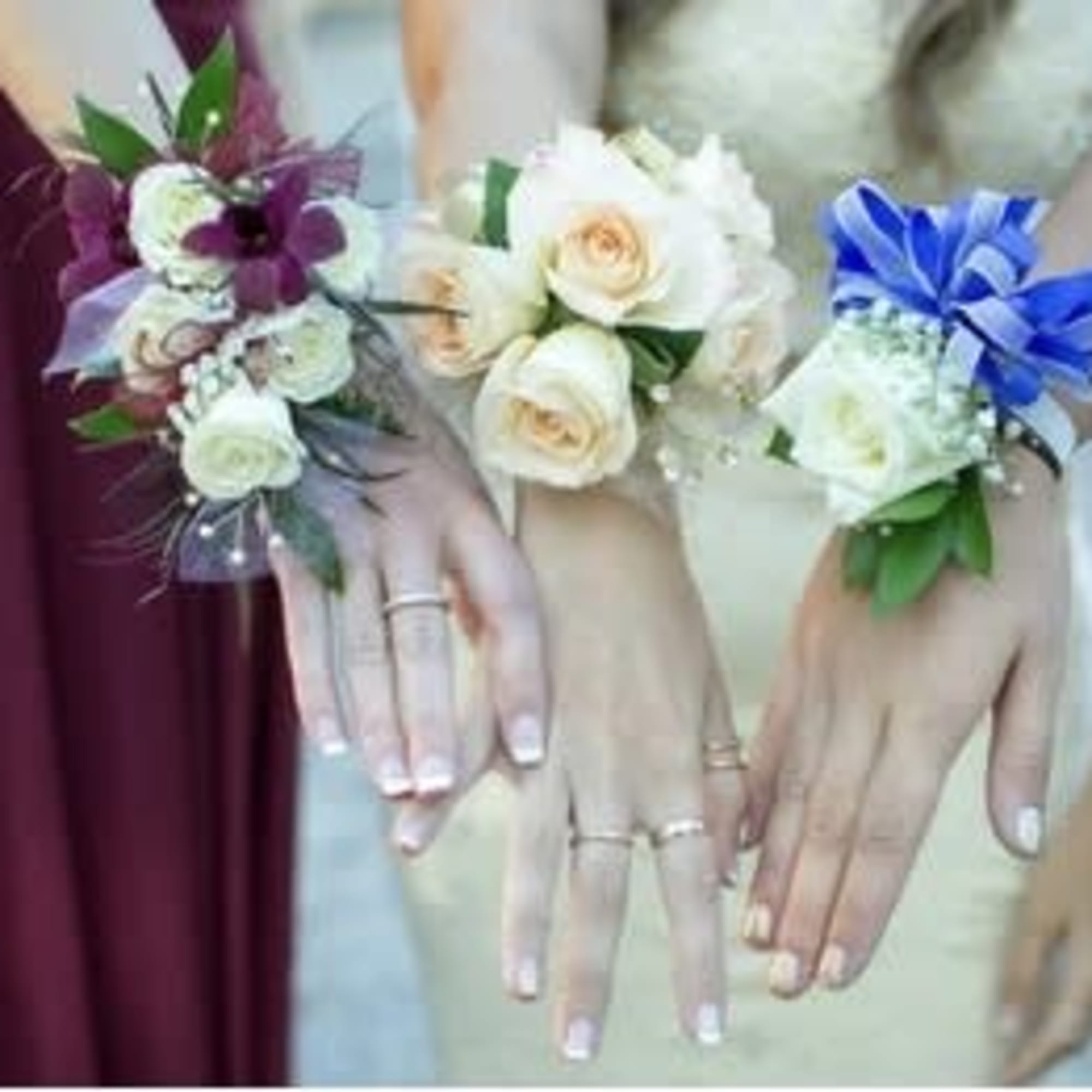 Three floral wrist corsages on women’s hands, in purple, white, and blue.