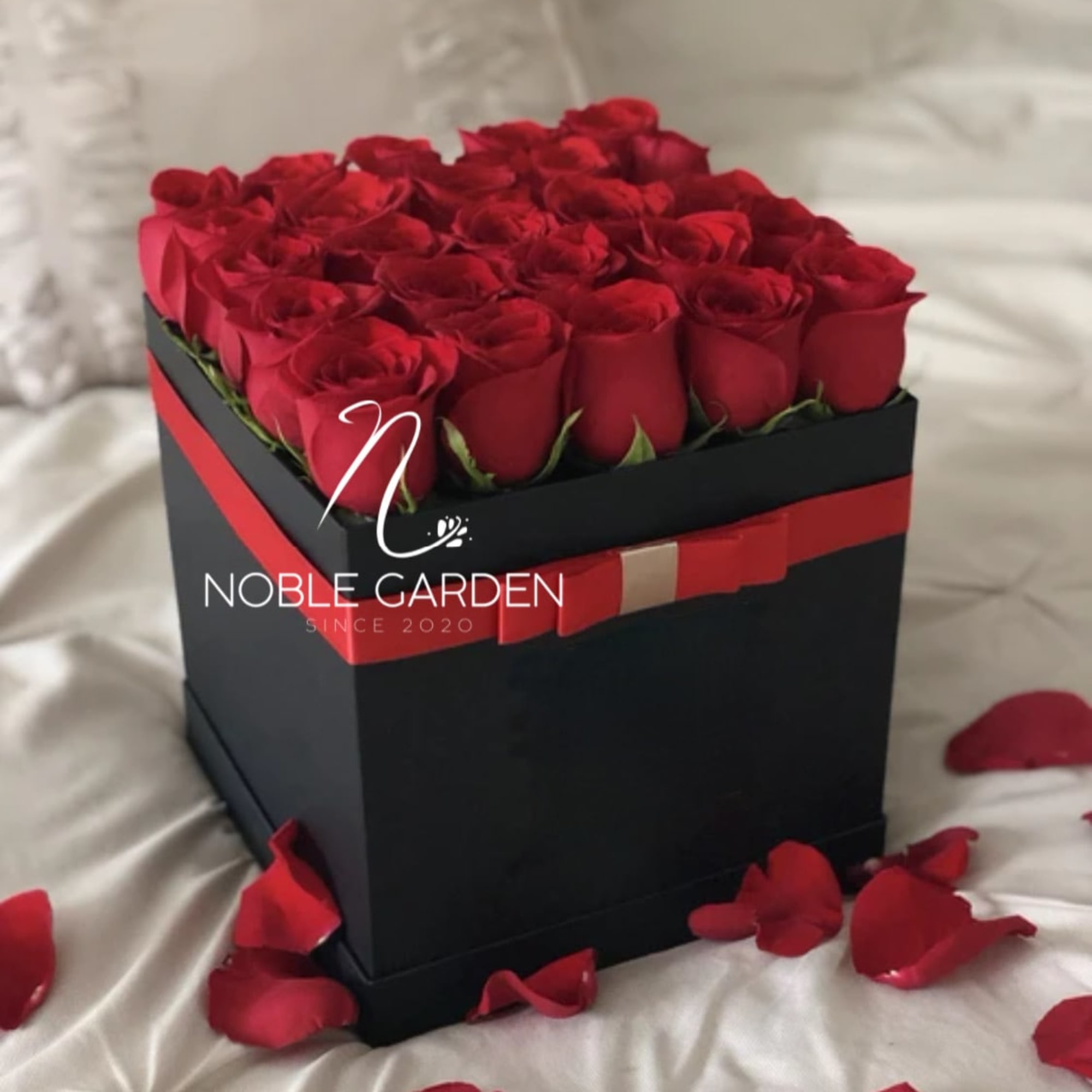 Red roses arranged in a black square gift box with rose petals around it
