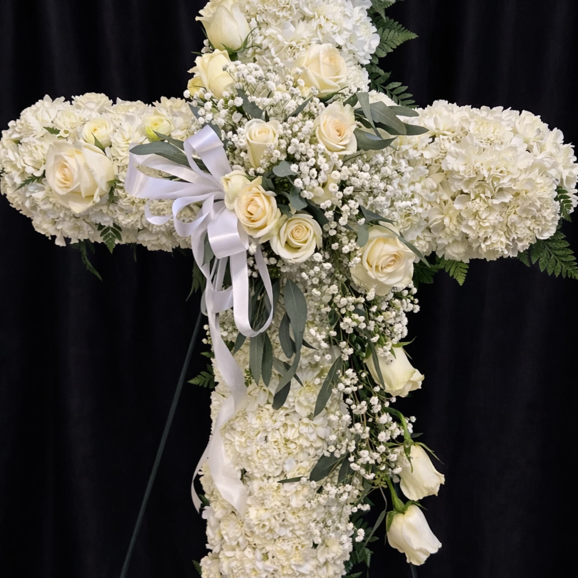 Tall white floral cross on an easel with roses and a white ribbon bow.