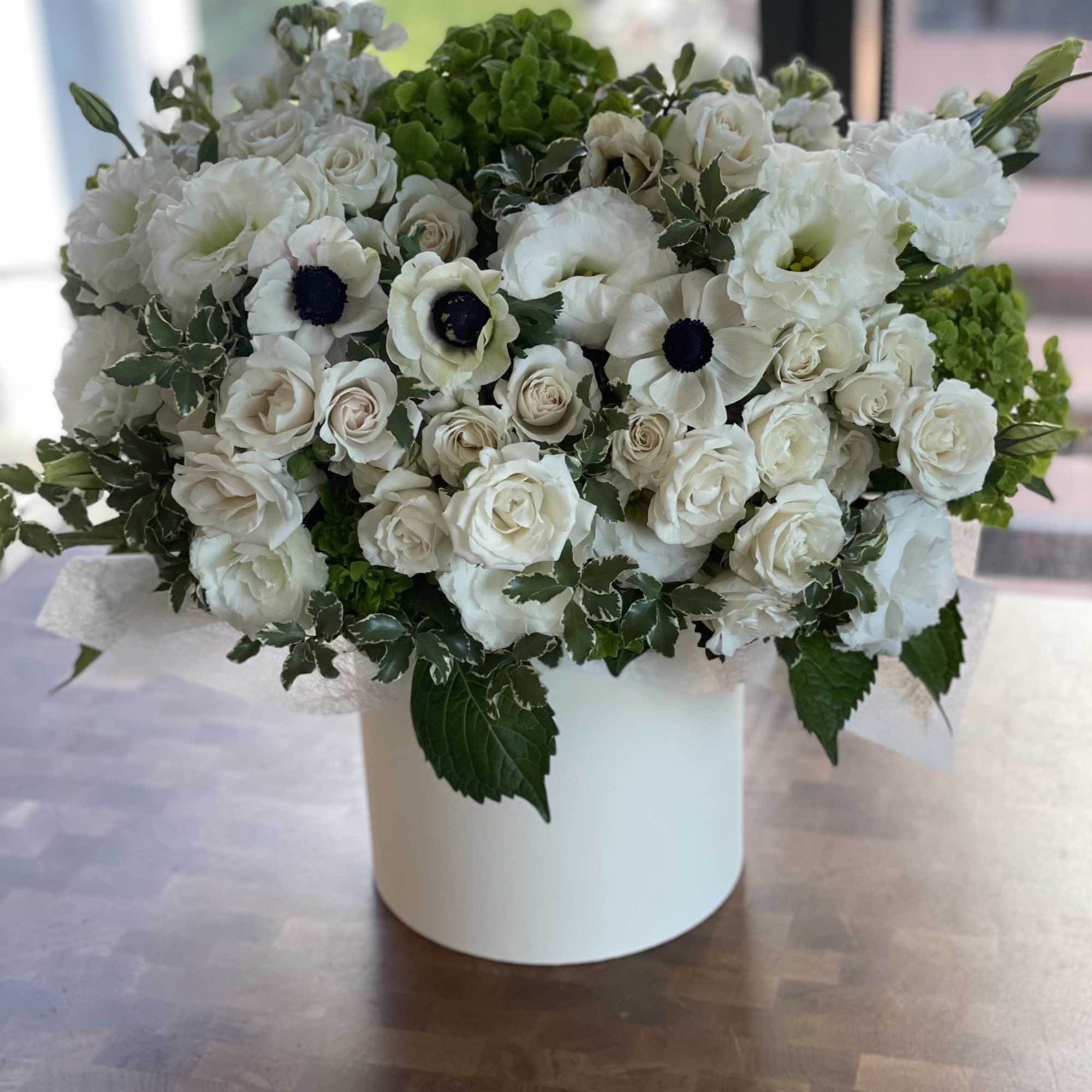Round arrangement of white roses, lisianthus, and anemones with green accents in a white cylinder vase