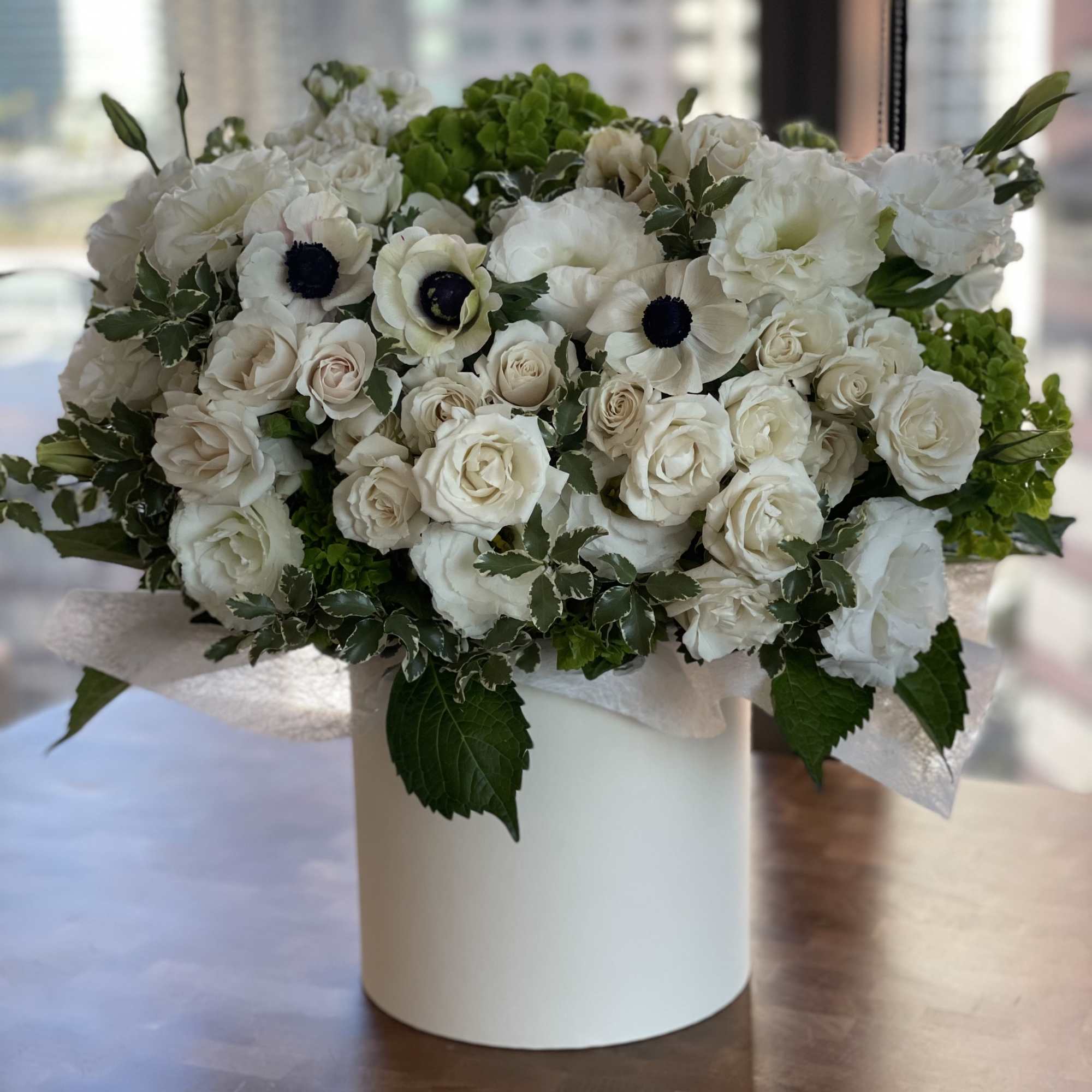 Lush white arrangement of roses and anemones with green accents in a round white hat box container