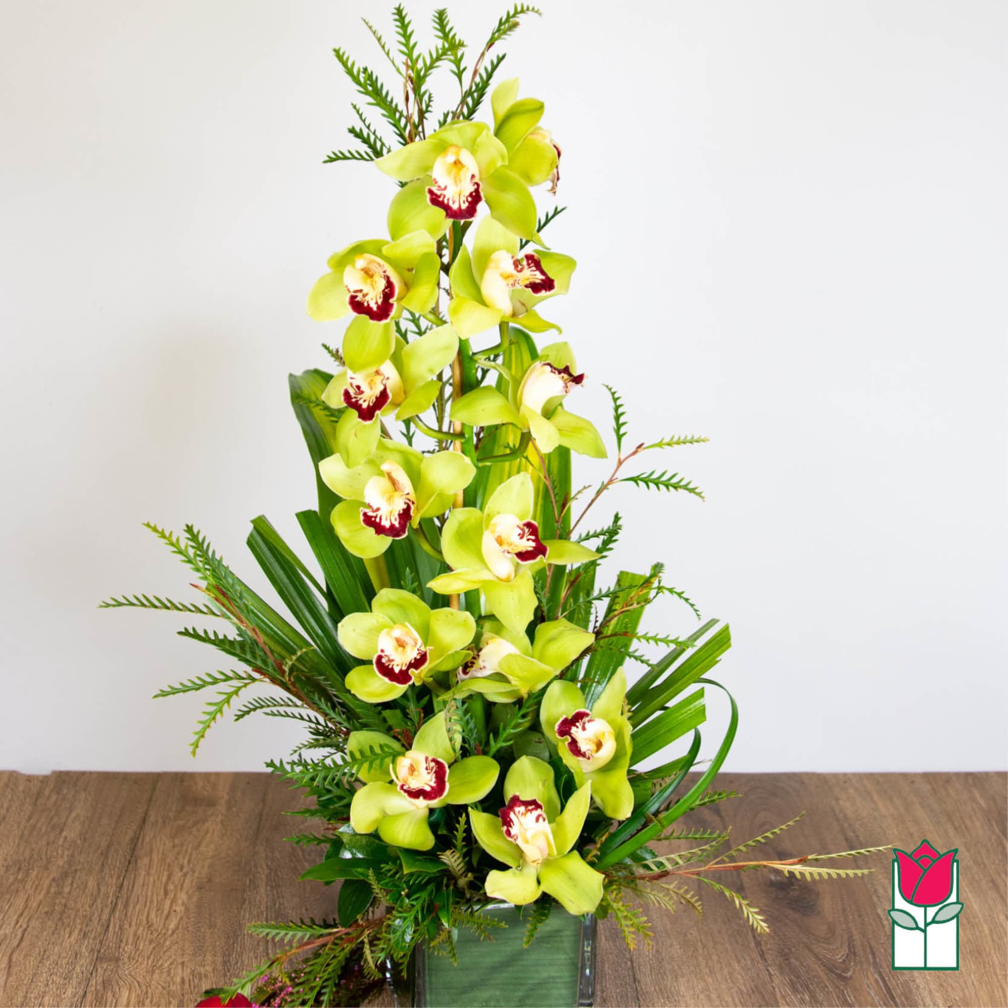 Tall arrangement of green cymbidium orchids with red centers in a green glass cube vase