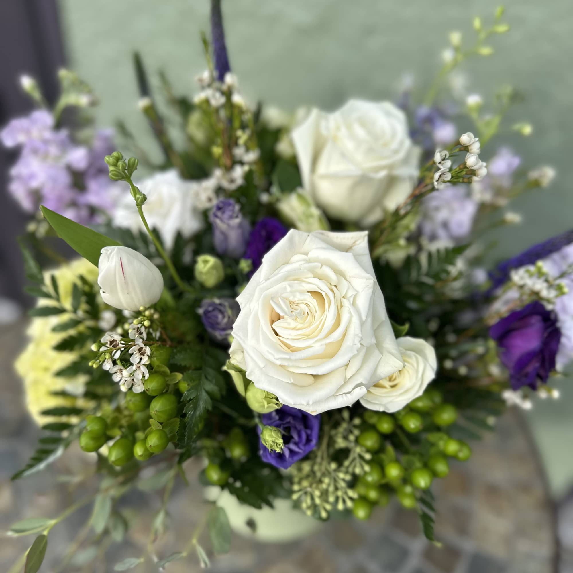 Arrangement of white roses and purple flowers with green berries in a pale vase.