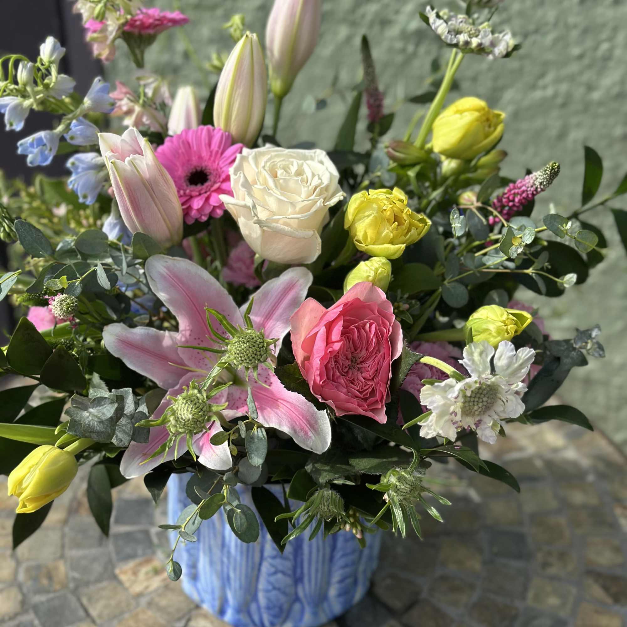 Mixed arrangement of pink lilies, roses, tulips and yellow blooms in a blue ceramic vase