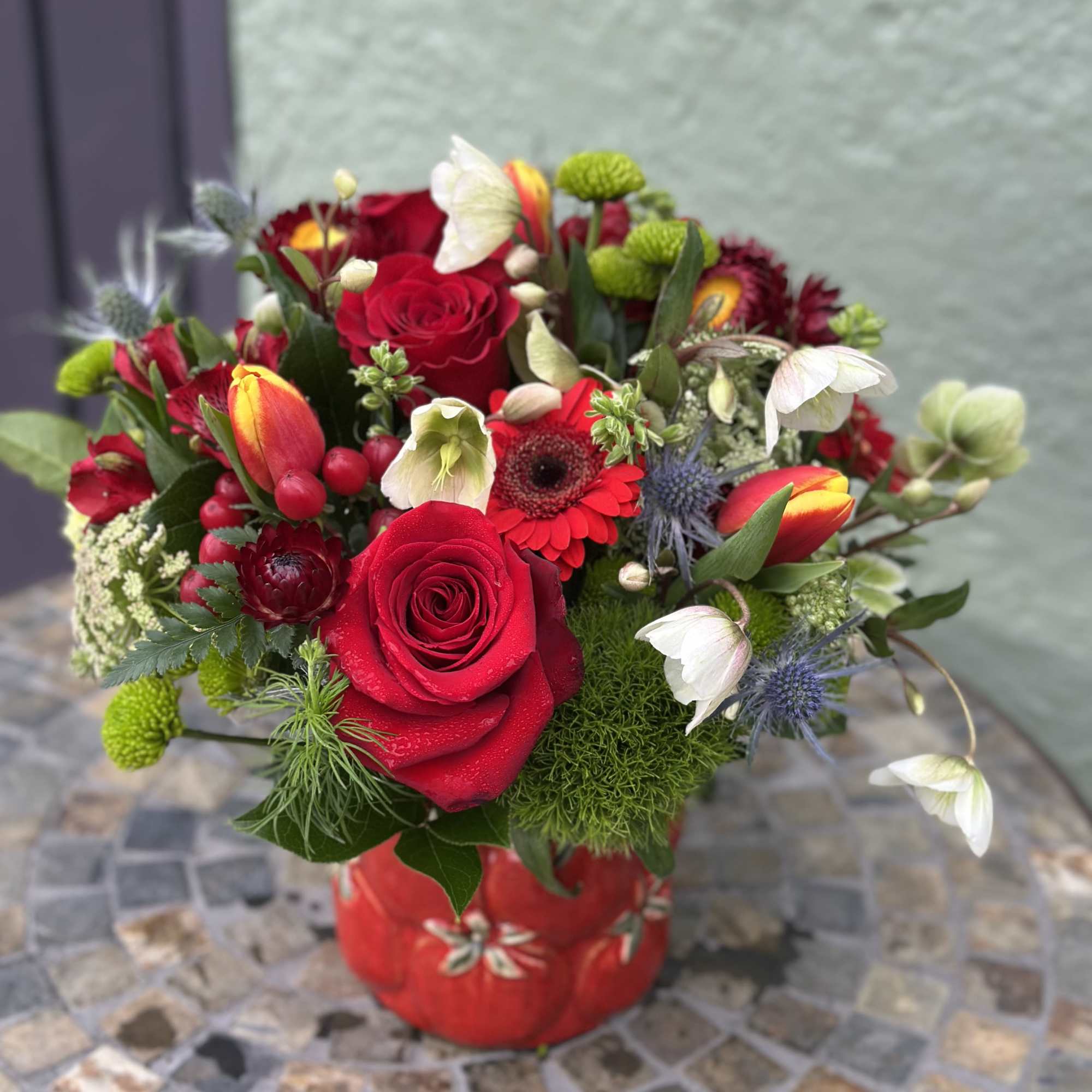 Compact arrangement of red roses, tulips, and mixed blooms in a red ceramic vase on a mosaic table.