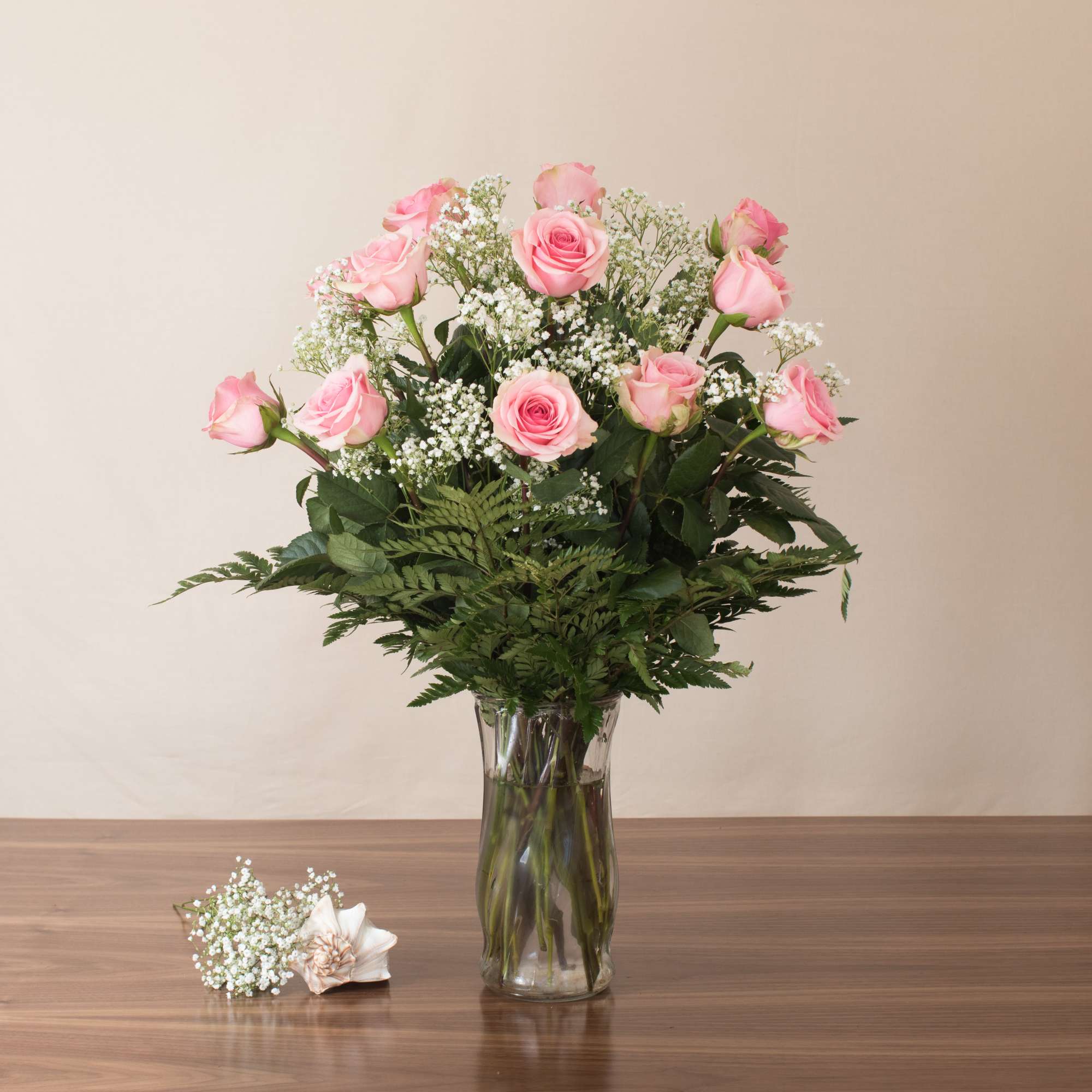 Tall glass vase of light pink roses with white filler flowers and a small matching corsage on the table.