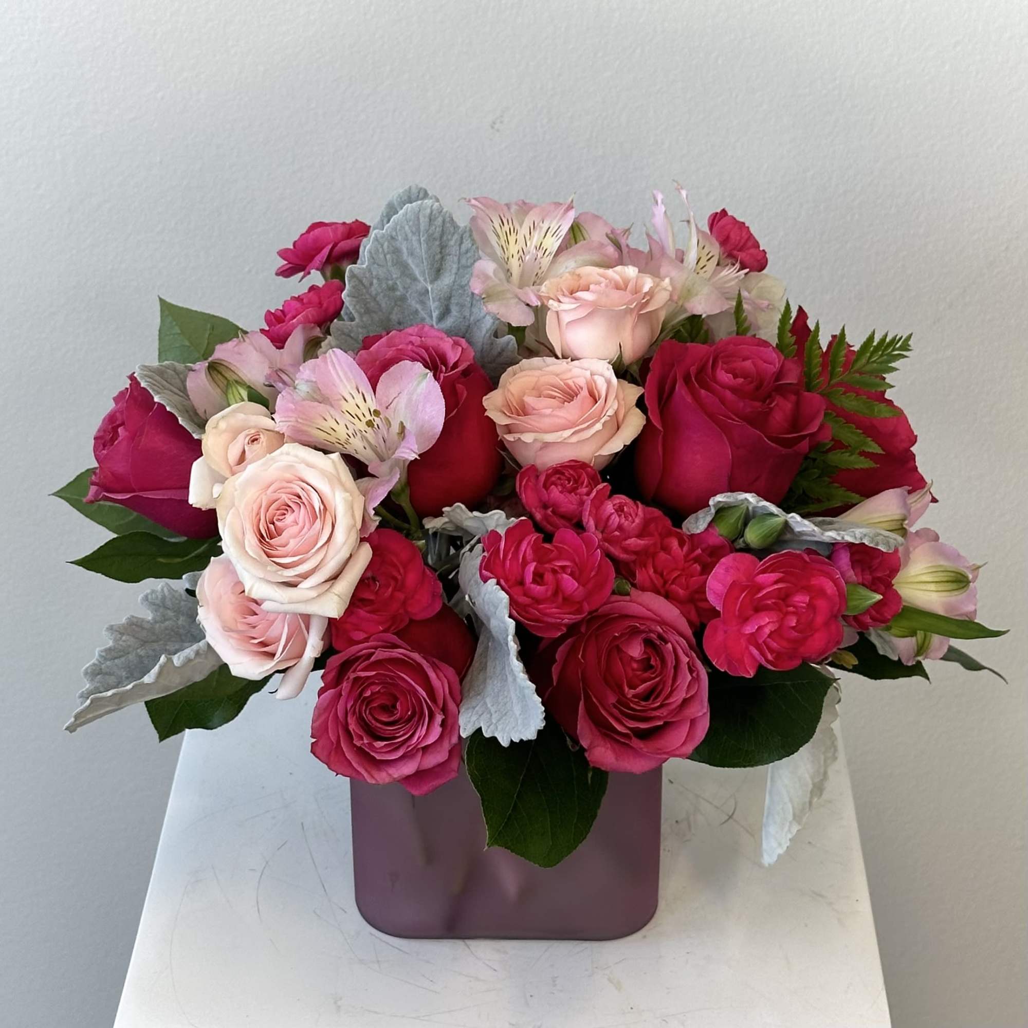 Compact arrangement of hot pink and blush roses with pink alstroemeria in a mauve cube vase