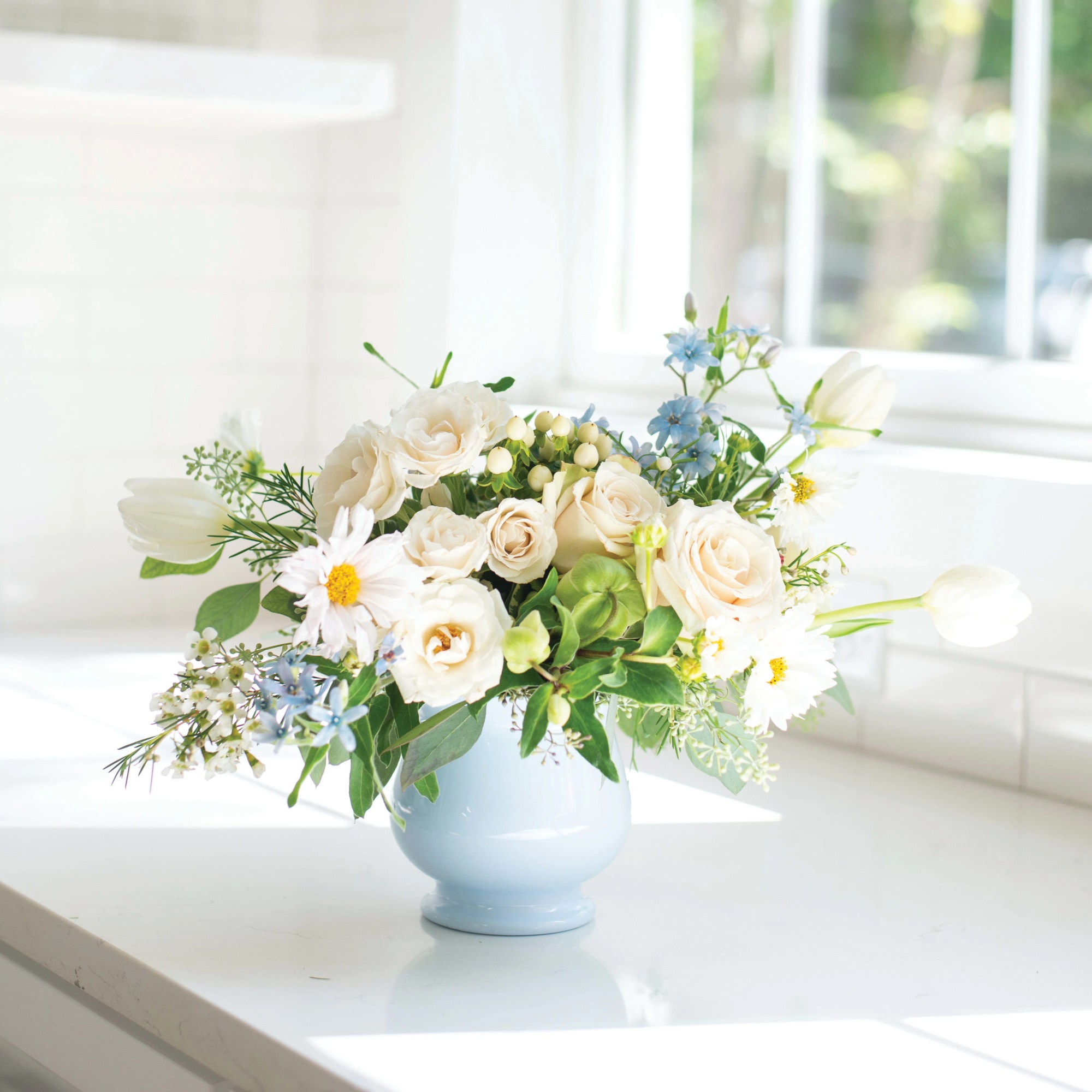 Low arrangement of cream roses, white tulips, and light blue blossoms in a pale blue vase on a sunlit counter