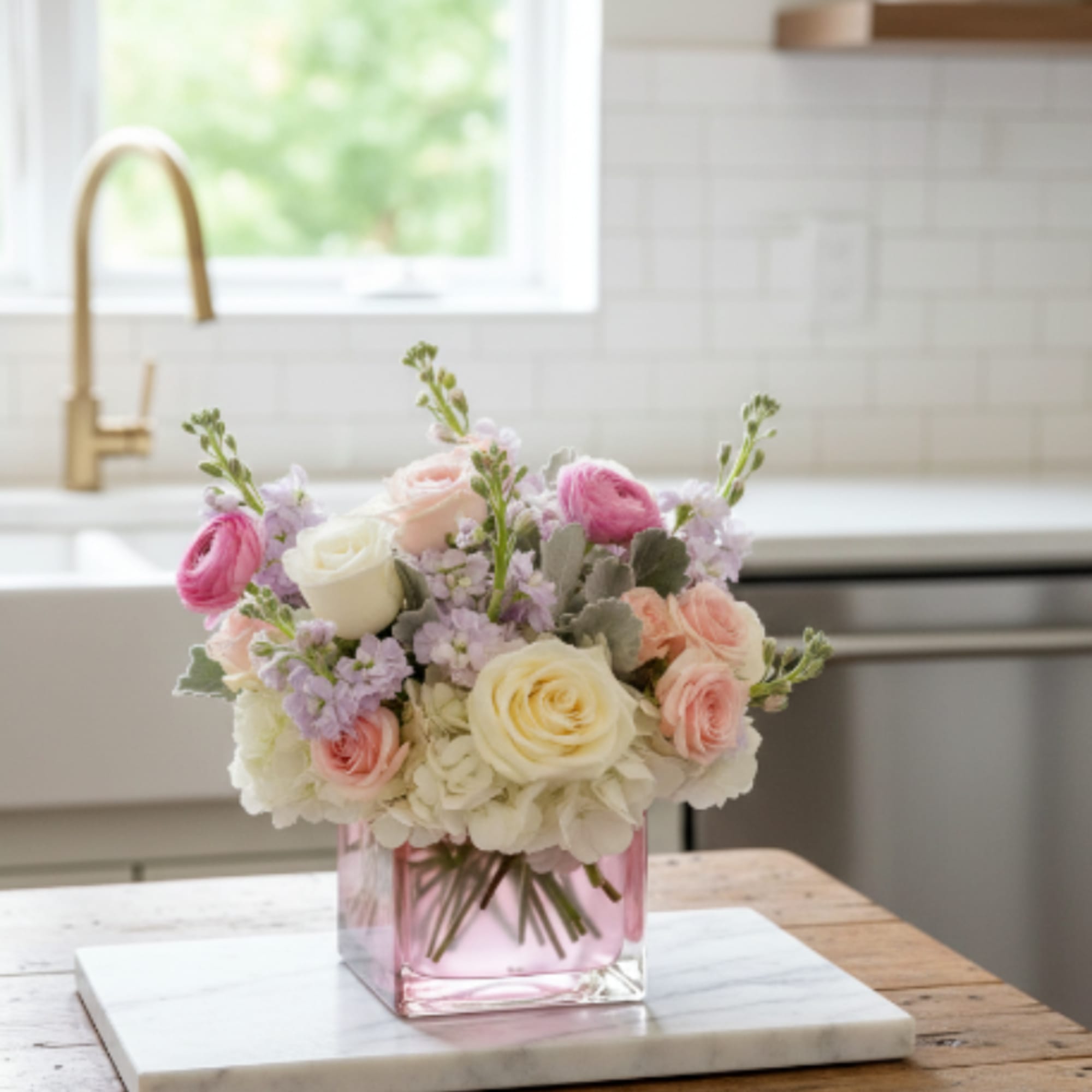 Pastel roses and ranunculus arranged in a pink glass cube vase on a wooden table.