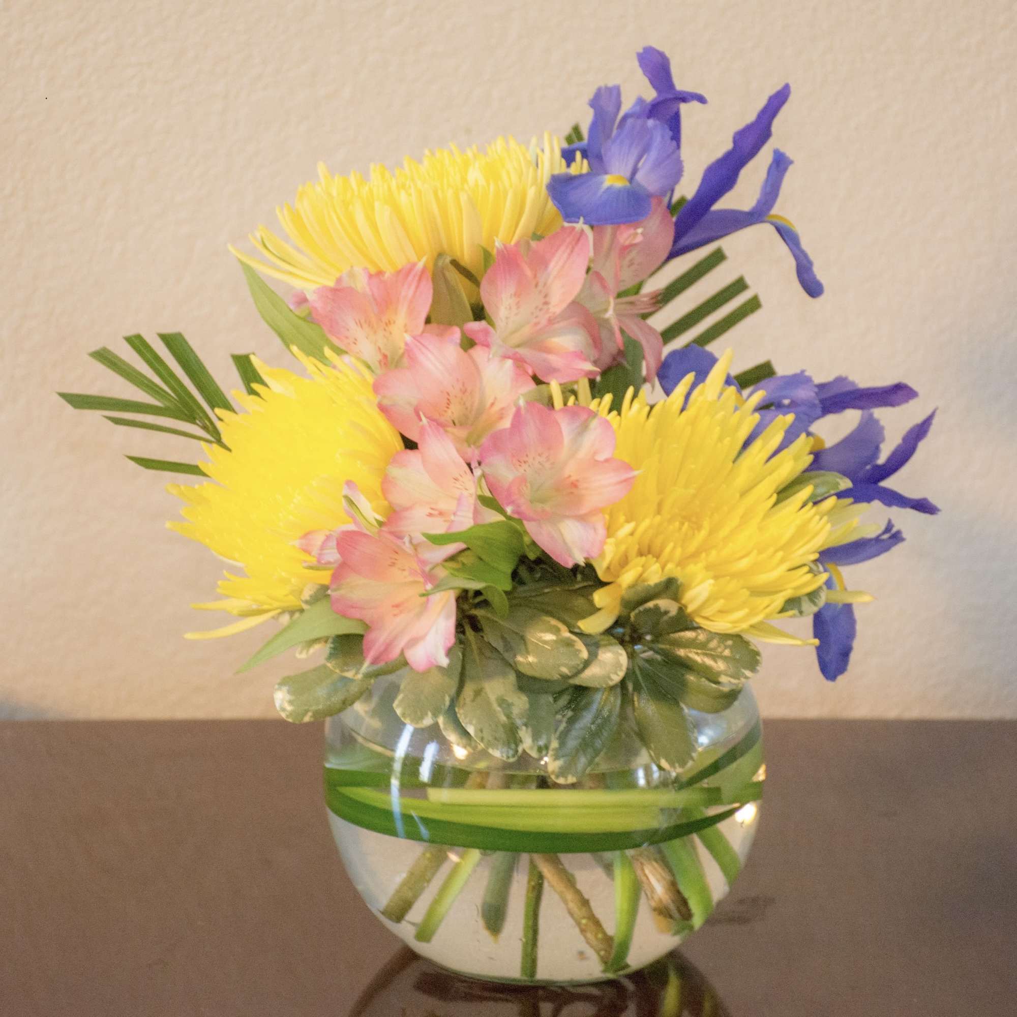 Small arrangement of yellow, pink, and blue flowers in a round glass vase with a green leaf wrap.