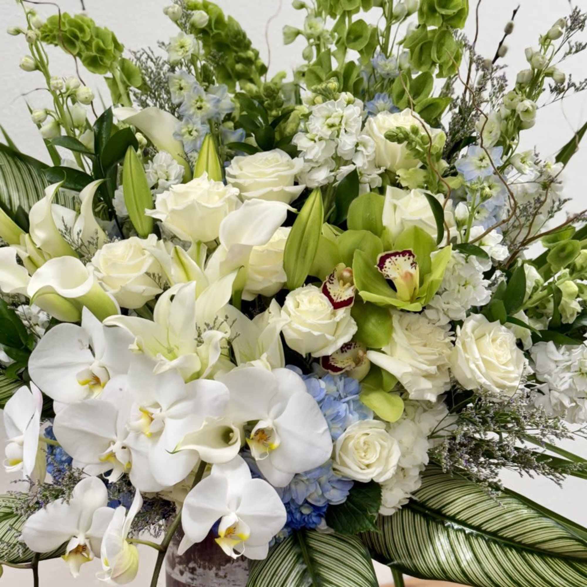 Tall white and green floral arrangement with orchids, roses, lilies, and large striped leaves in a rustic vase.