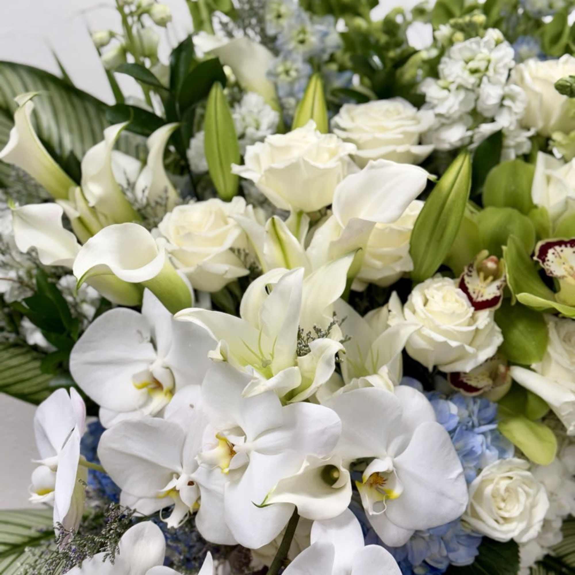 Tall white arrangement with roses, orchids, lilies, calla lilies, blue hydrangea, and green cymbidium orchids.