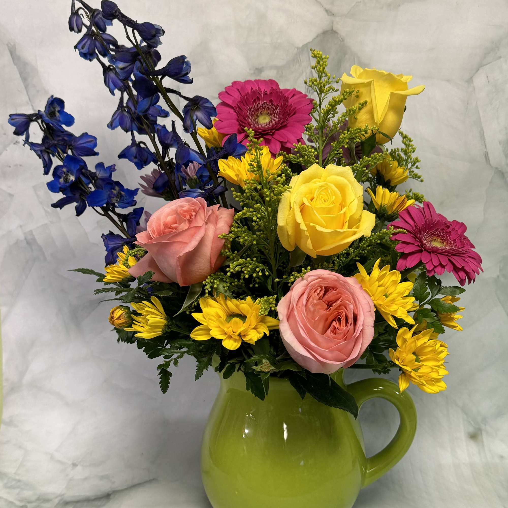 Colorful bouquet of roses, gerbera daisies, and blue delphinium in a green pitcher vase