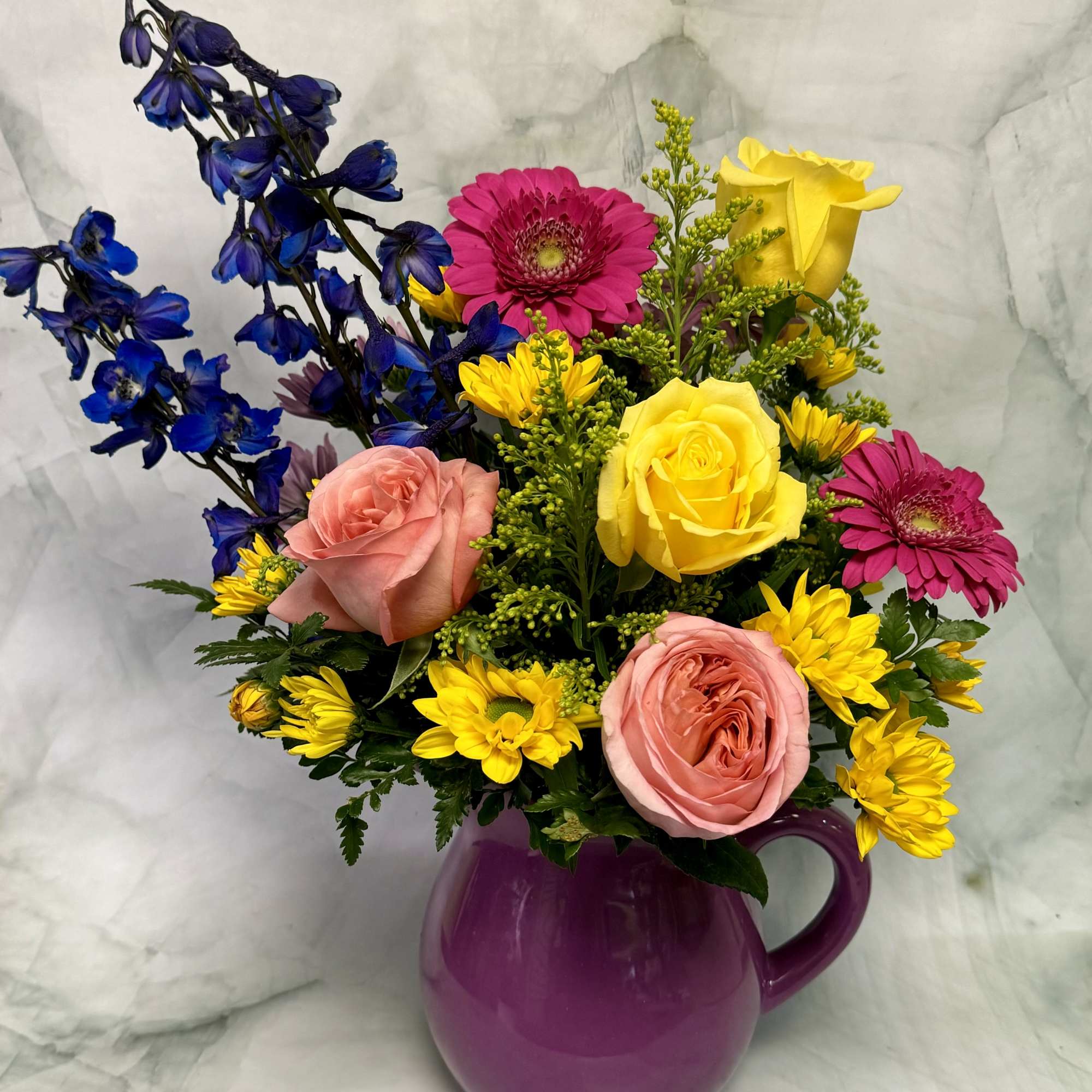Mixed bouquet in a purple pitcher with roses, gerbera daisies, and blue delphinium