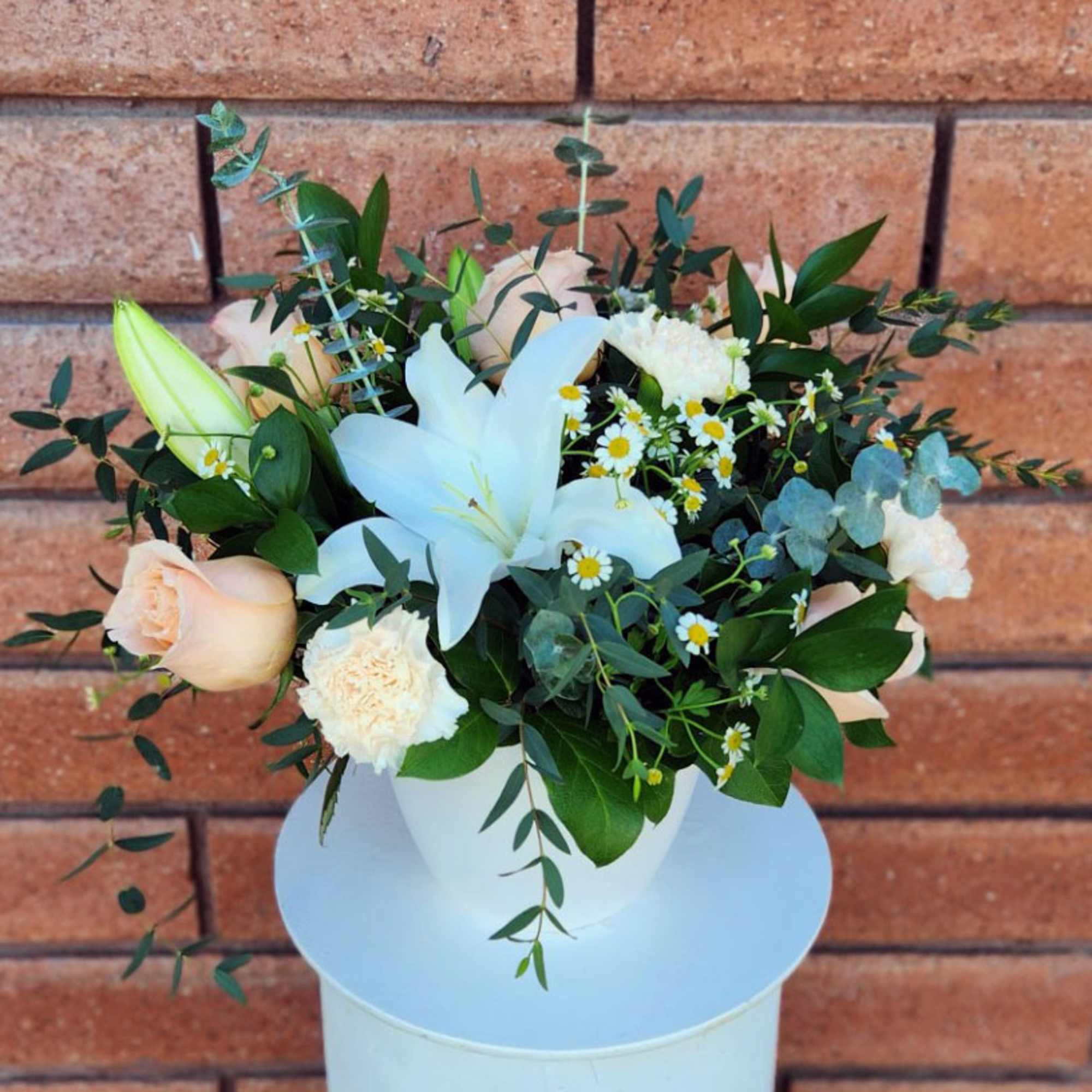 White lilies, peach roses, carnations, and small daisies in a white pot against a brick wall.