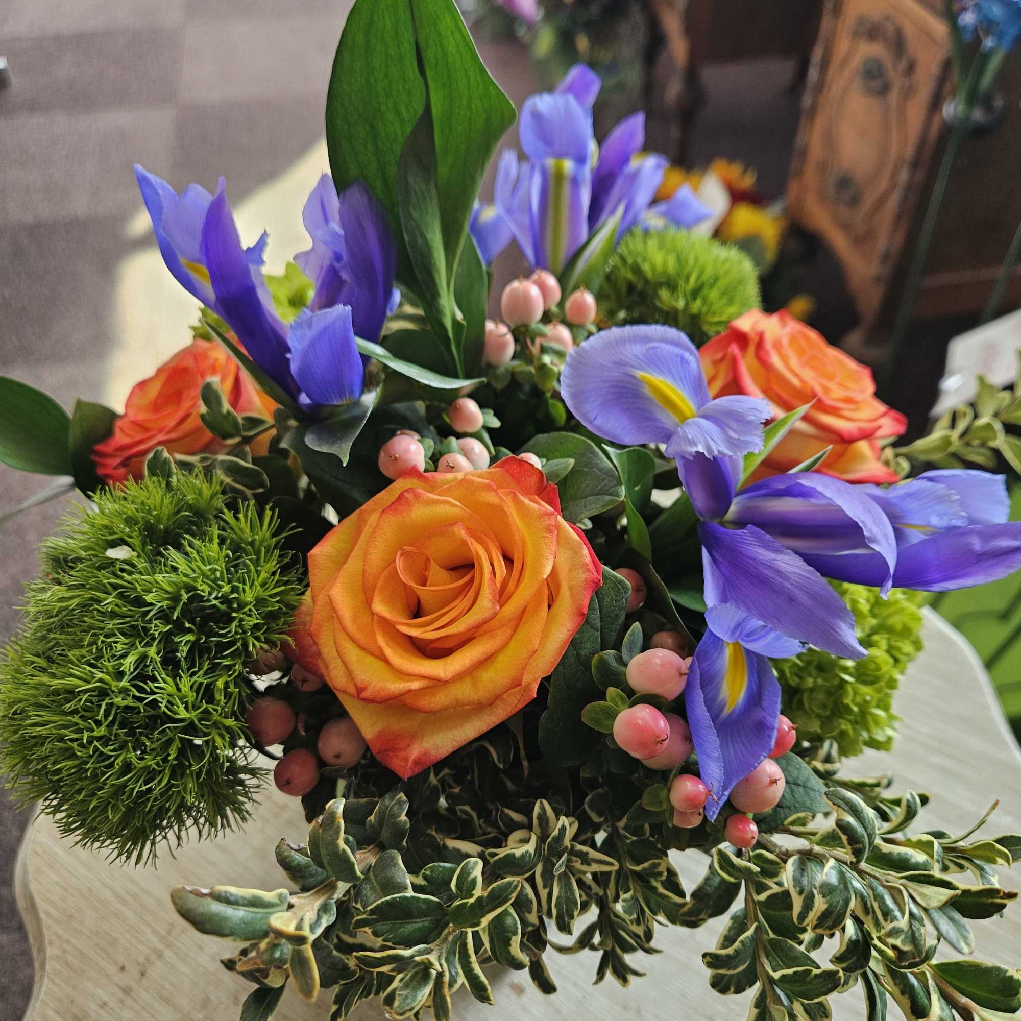 Low arrangement of orange roses, purple irises, green dianthus and pink berries in a compact container
