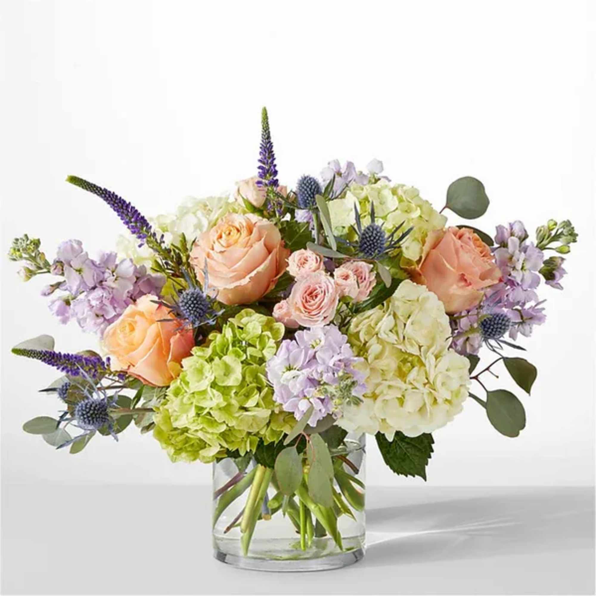 Arrangement of peach roses, green and ivory hydrangeas, and purple blooms in a clear glass vase
