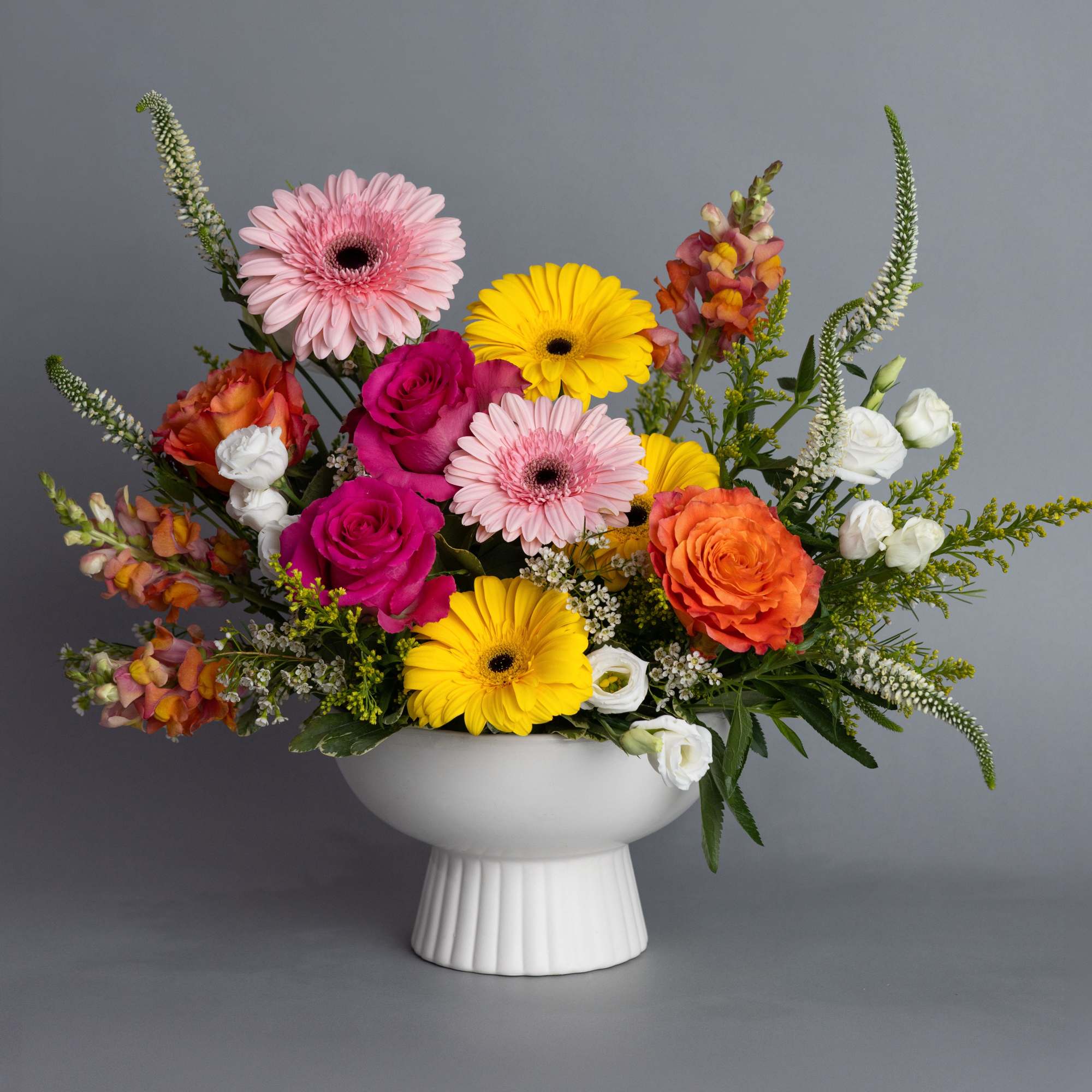 Bright mixed arrangement of gerbera daisies, roses, and lisianthus in a white pedestal bowl