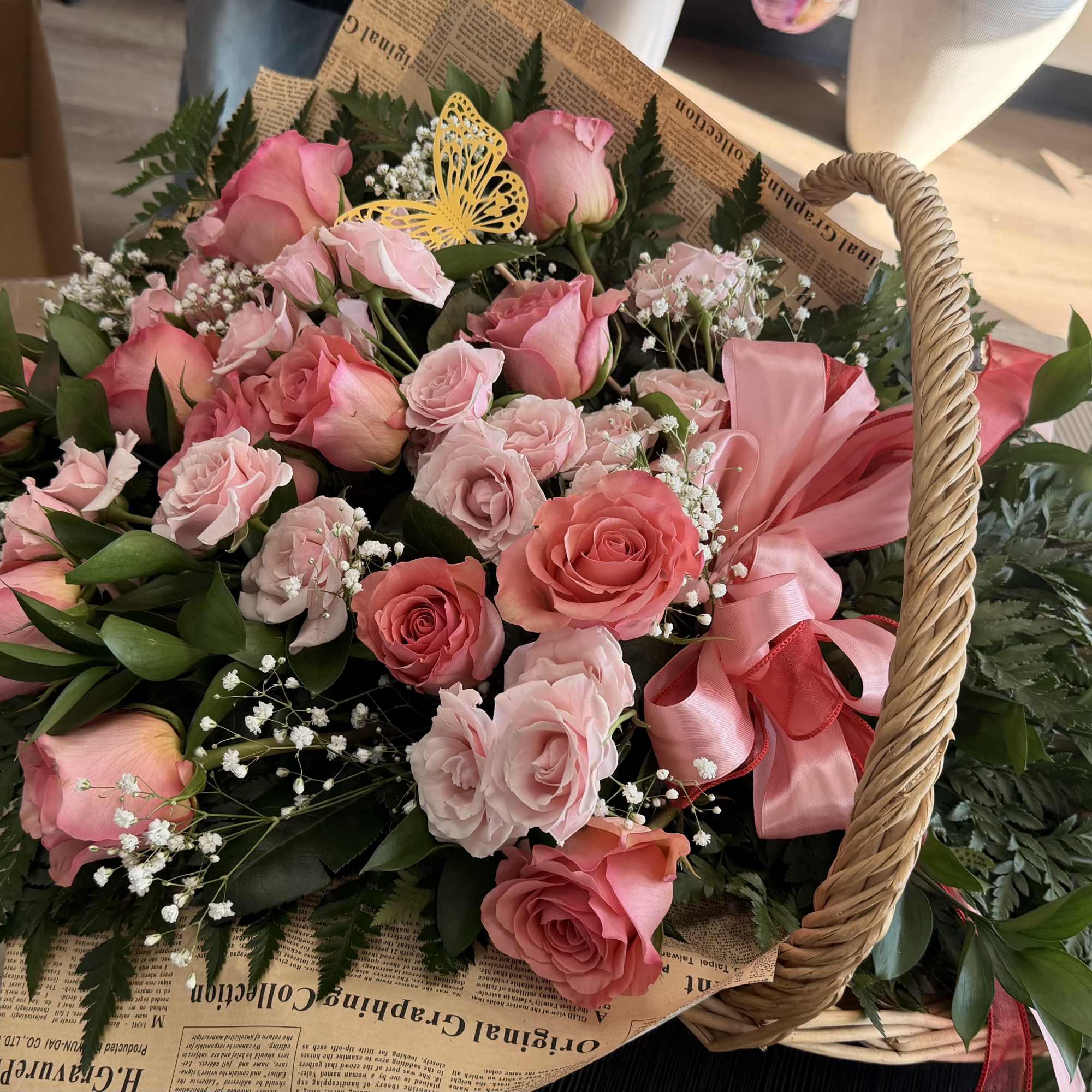 Basket arrangement of pink roses and spray roses with baby's breath, pink bow, and yellow butterfly accent