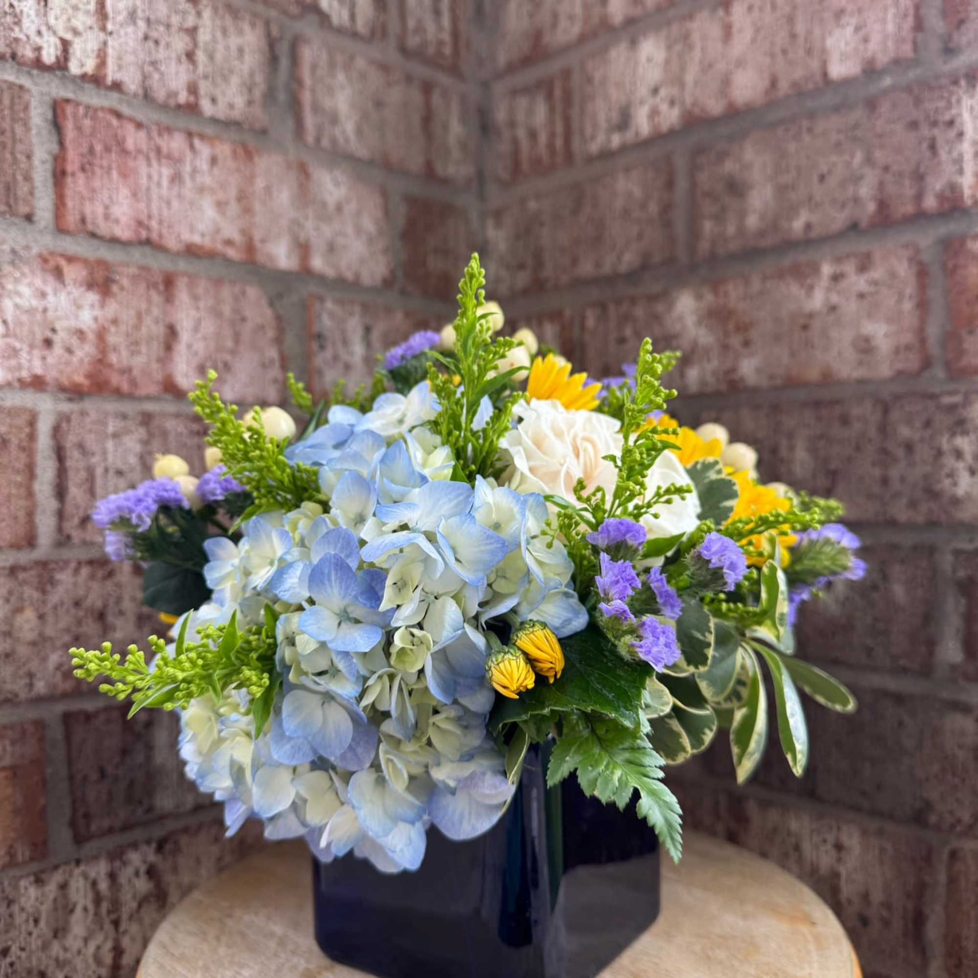 Blue hydrangea bouquet in a black square vase with yellow and purple accents