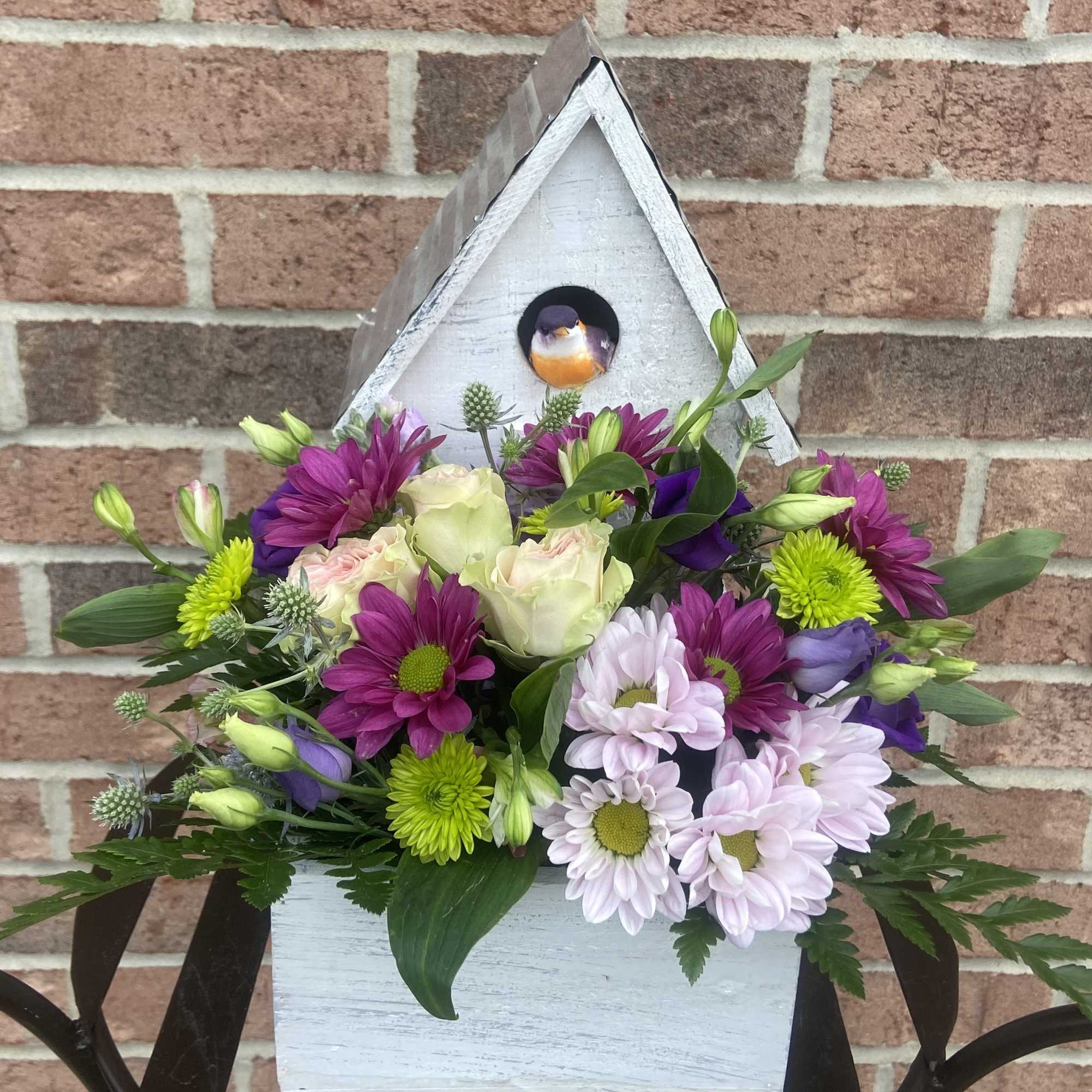 Mixed purple, white, and green flowers arranged in a white birdhouse container