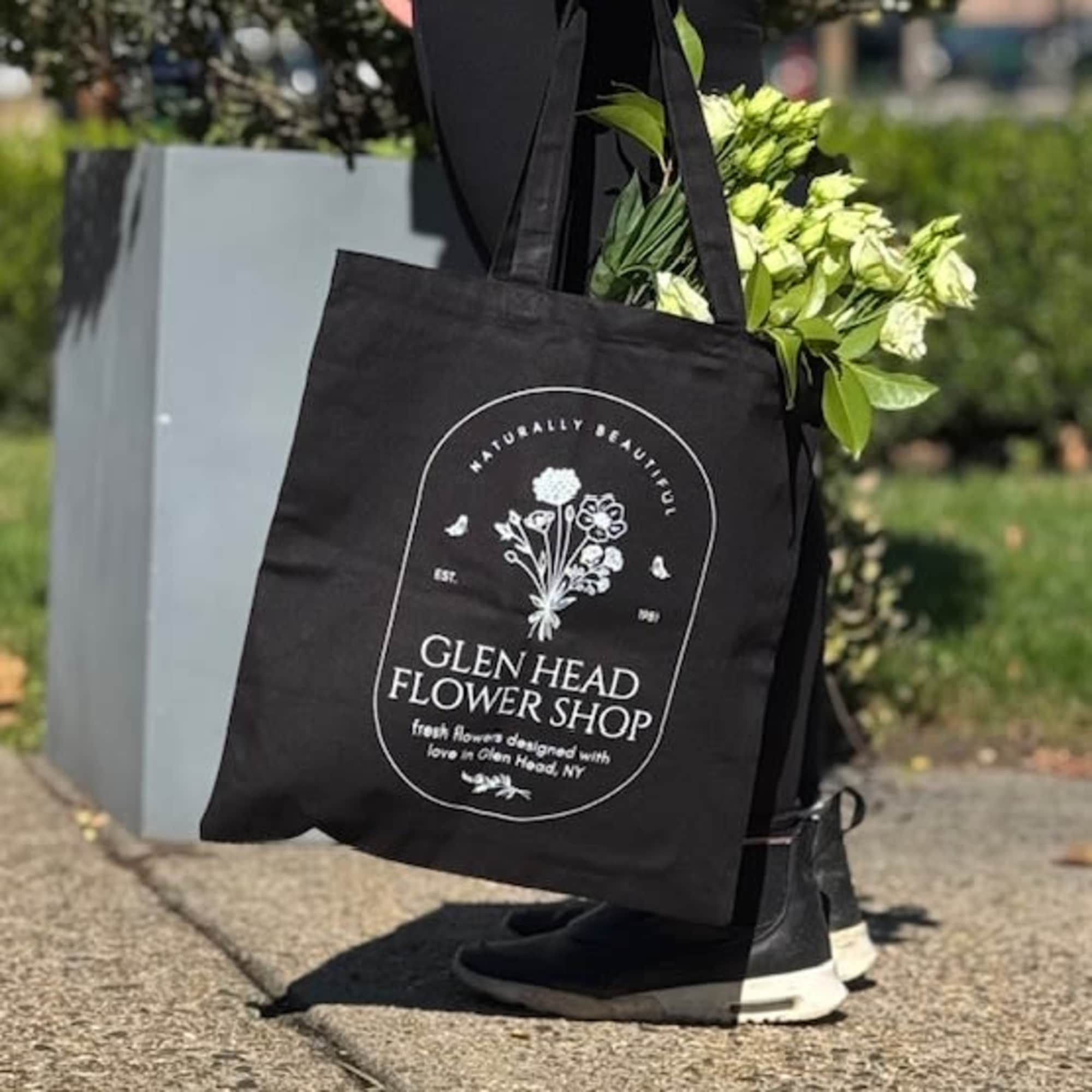 Black flower shop tote bag with white logo, filled with small white flowers, carried outdoors by a person.