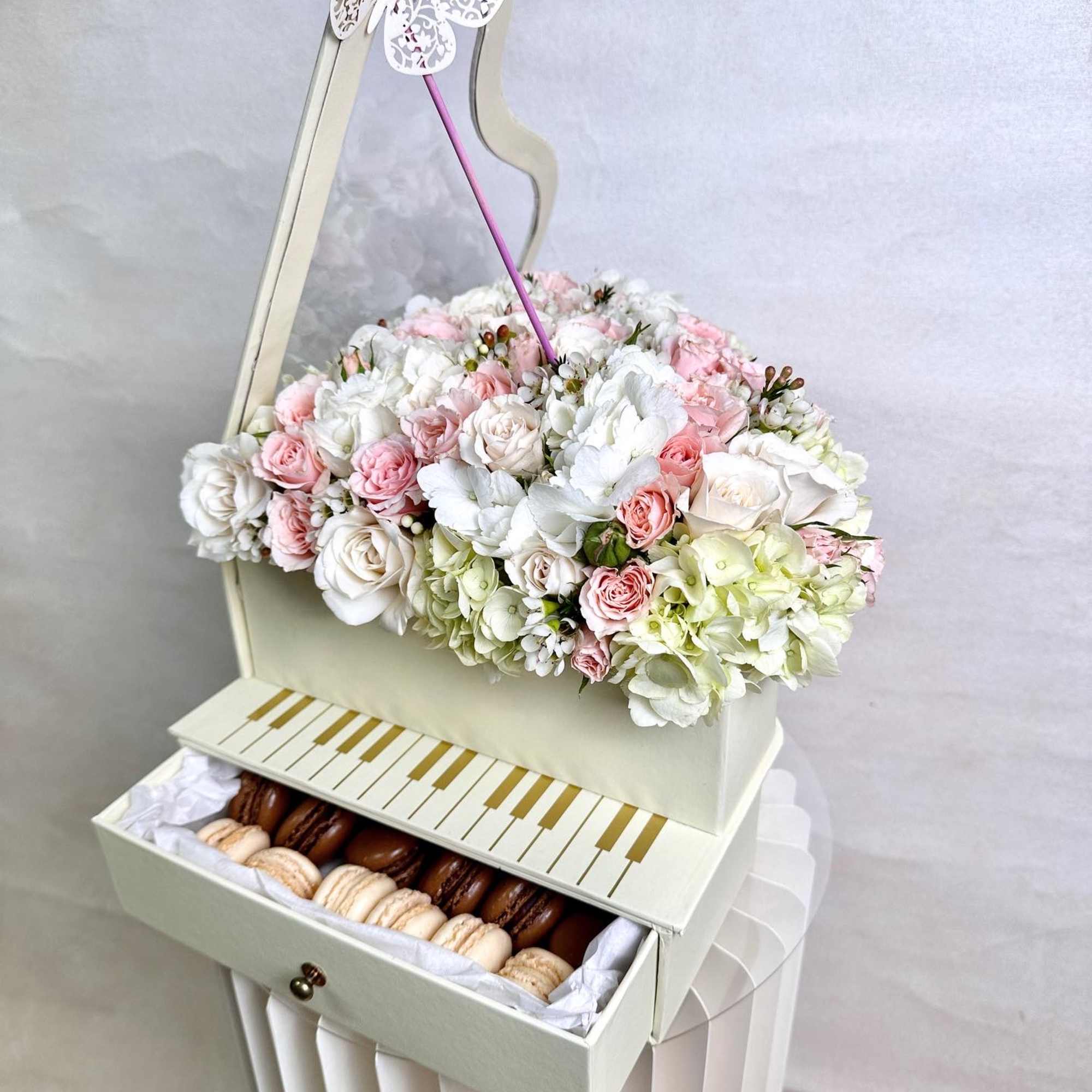 Piano-shaped cream box topped with pastel roses and hydrangeas, with a drawer of assorted macarons.