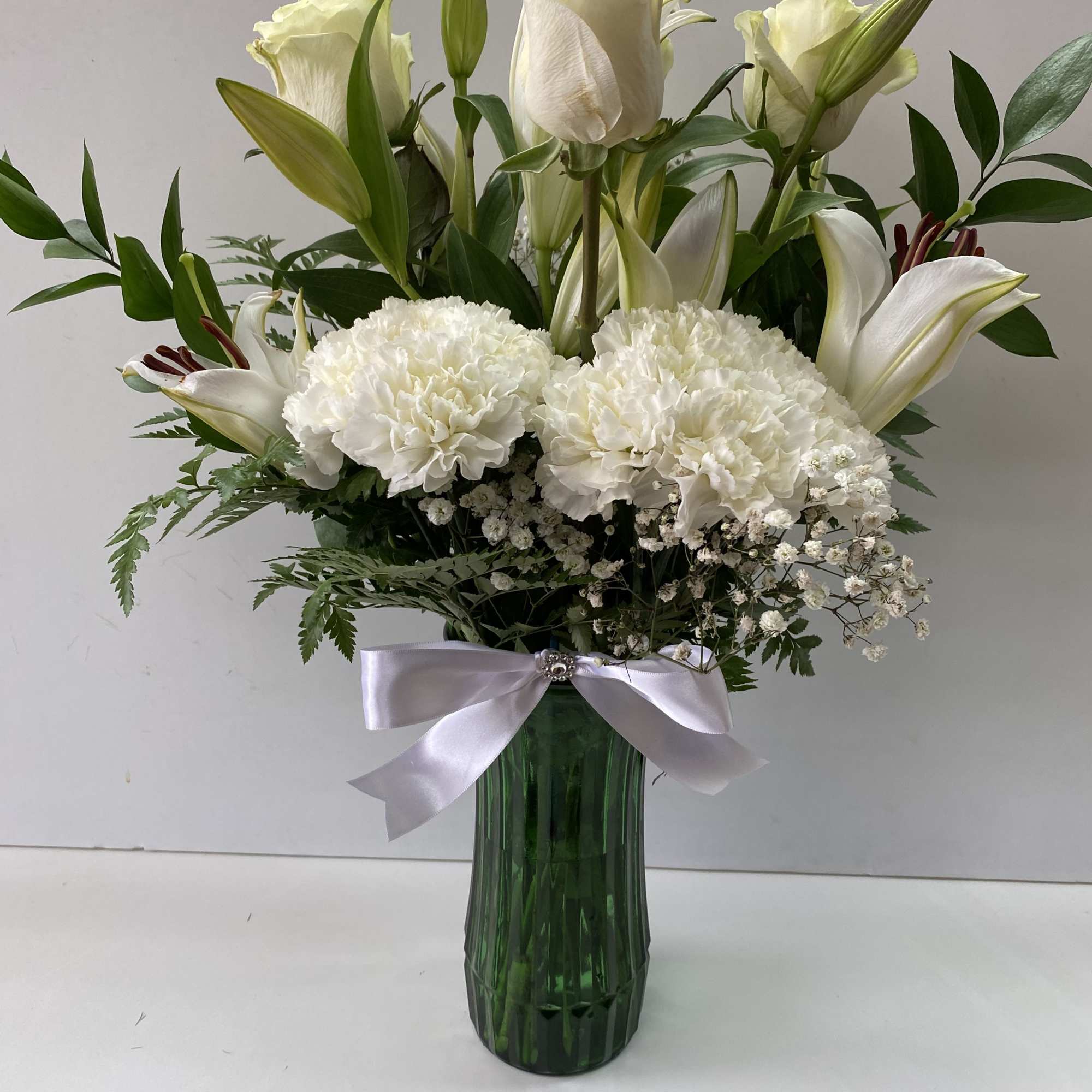Tall arrangement of white roses, lilies, and carnations in a green vase with white ribbon bow