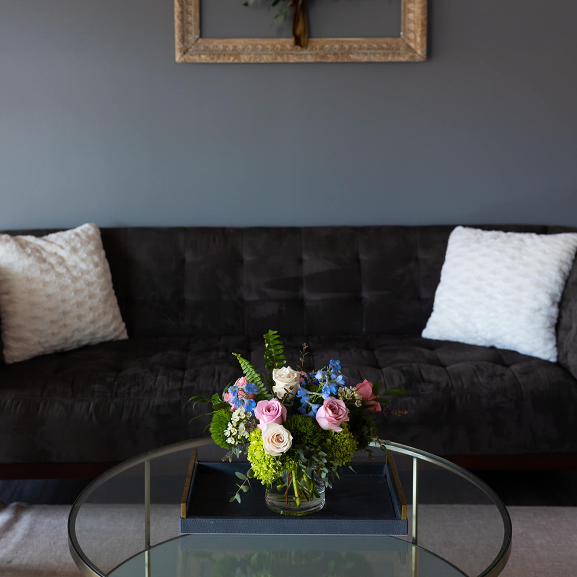 Low mixed arrangement of pink roses, blue flowers, and green blooms in a glass vase on a round glass coffee table