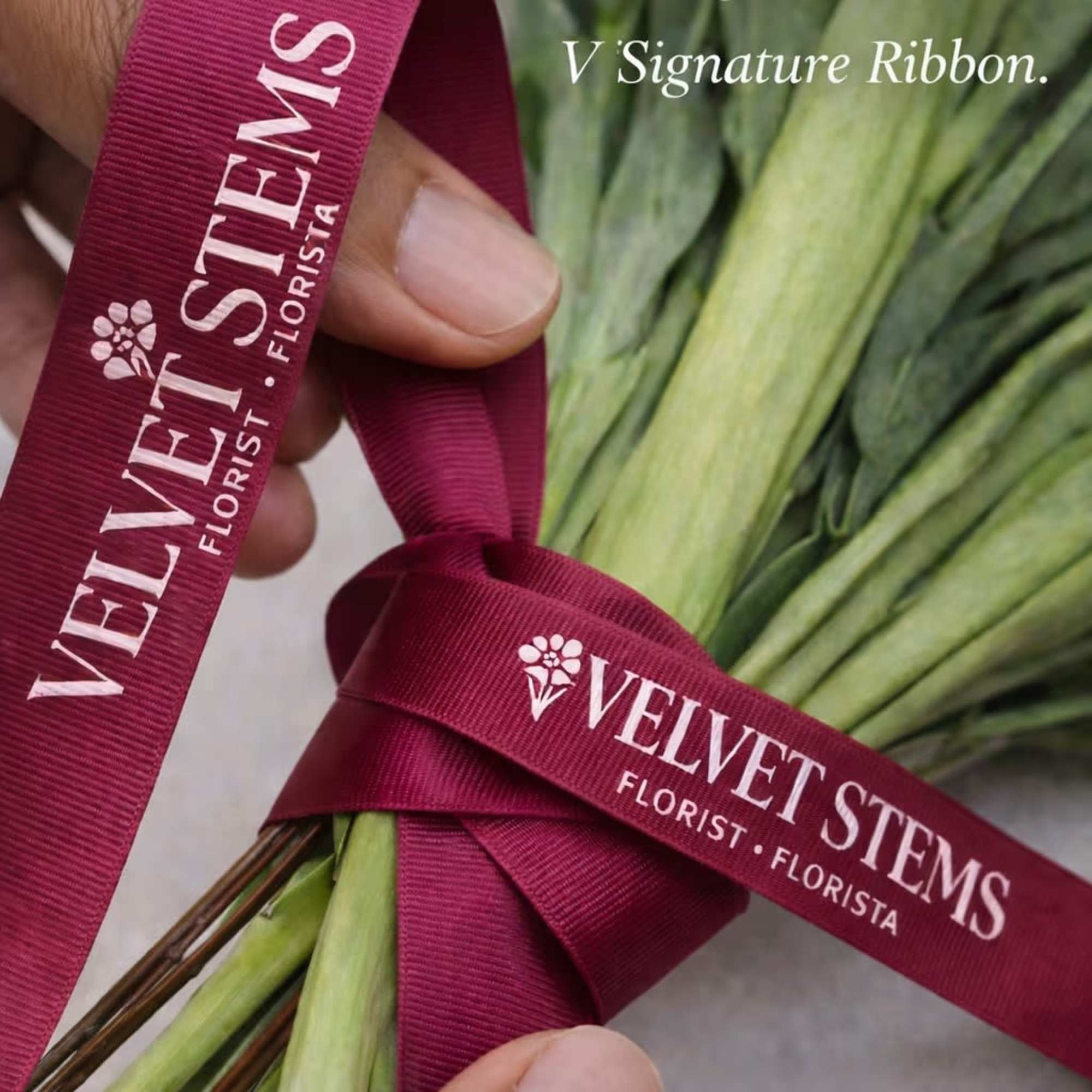 Hand tying a dark berry ribbon with florist logo around a bouquet’s stems