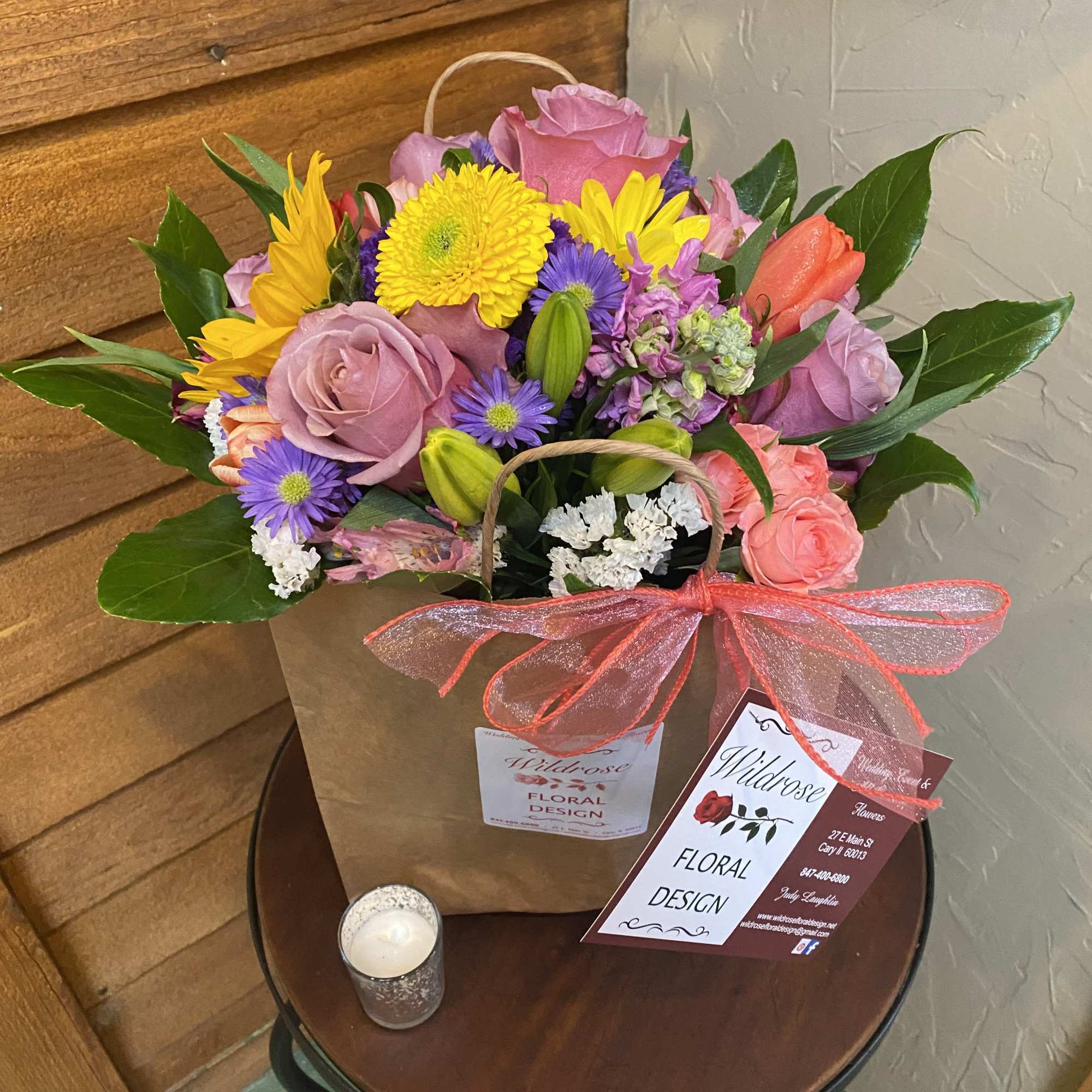Mixed bouquet of pink roses, yellow daisies, and purple flowers in a paper bag