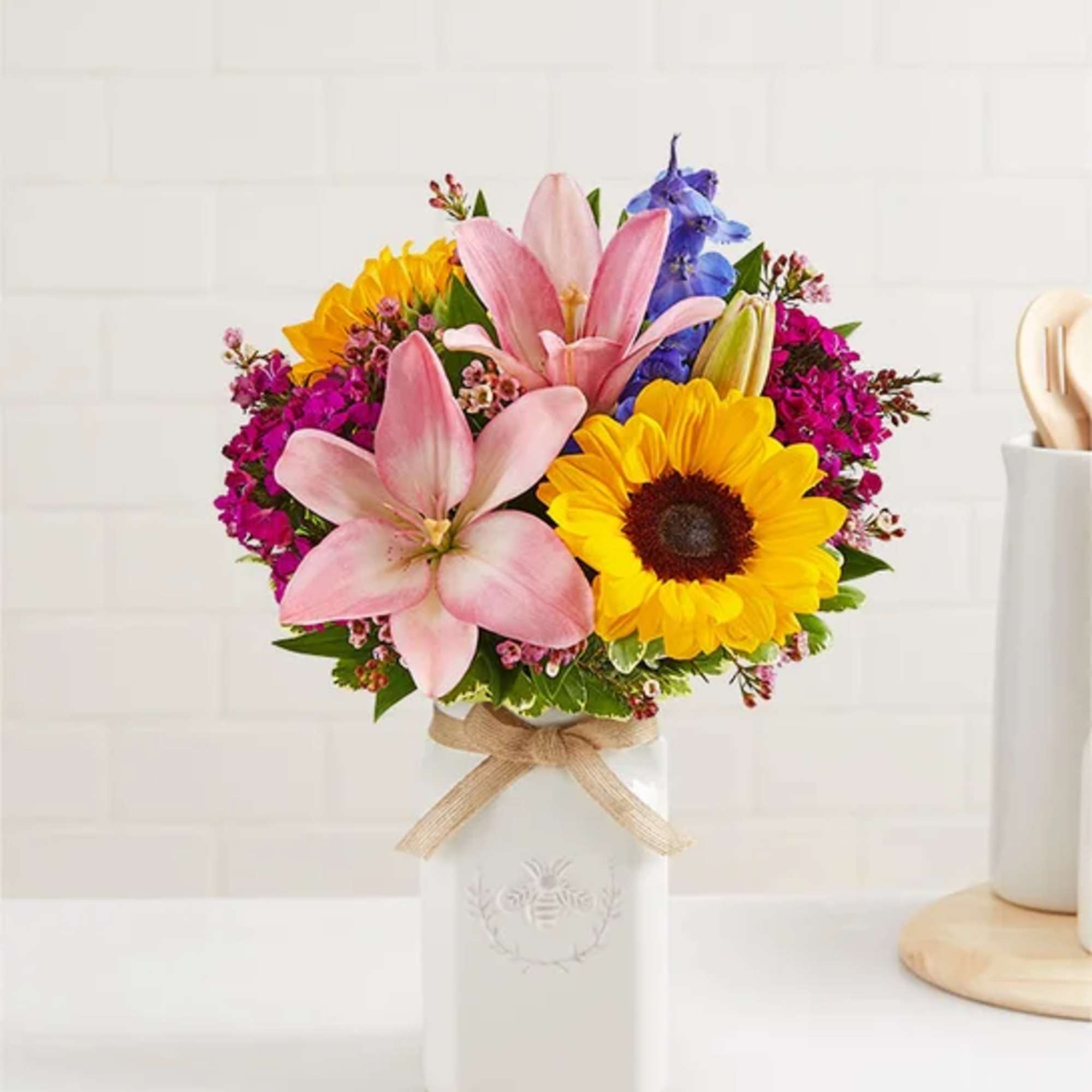Colorful bouquet of pink lilies, sunflowers and purple blooms in a white ceramic jar with twine bow.