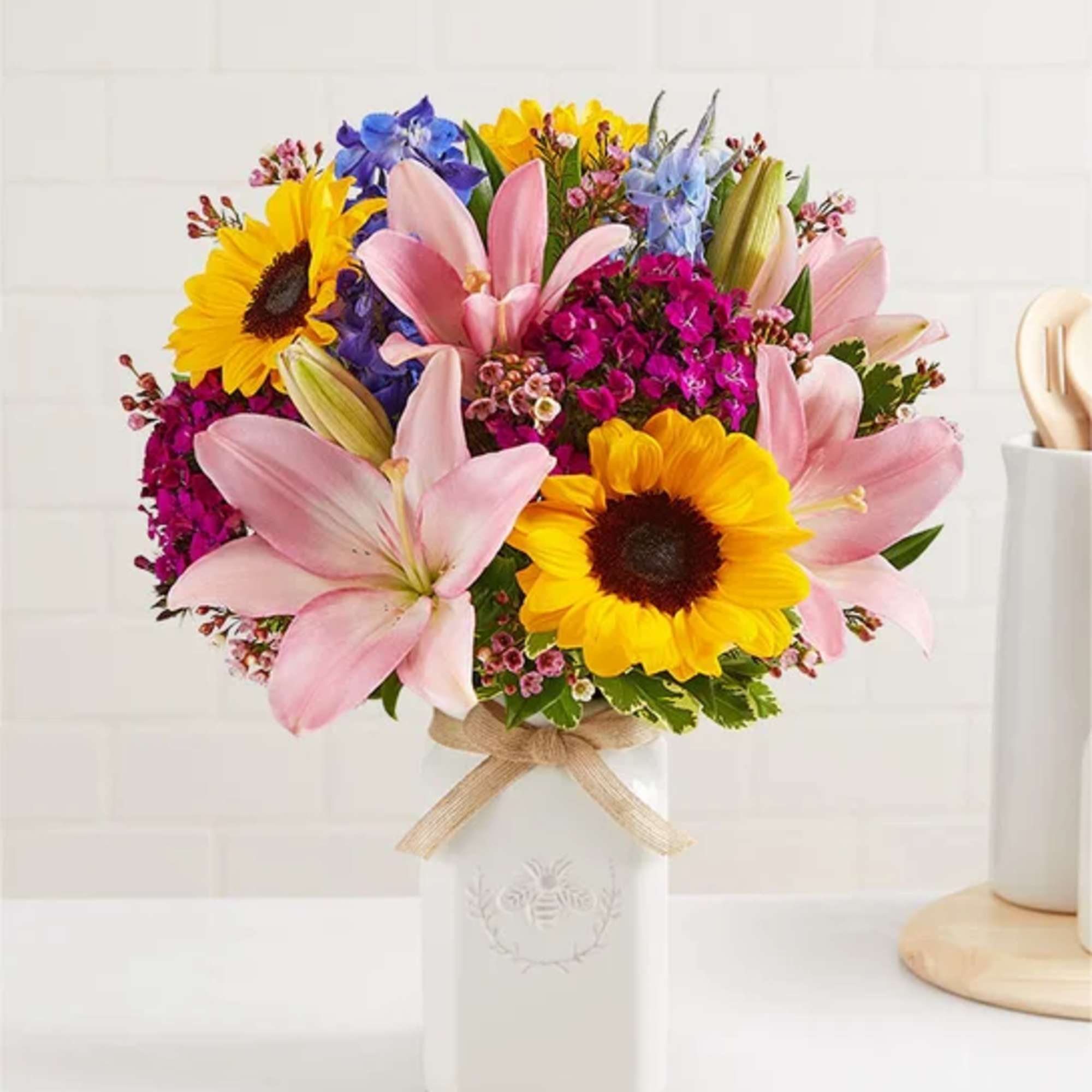 Bright bouquet of pink lilies, yellow sunflowers and mixed blooms in a white ceramic vase.