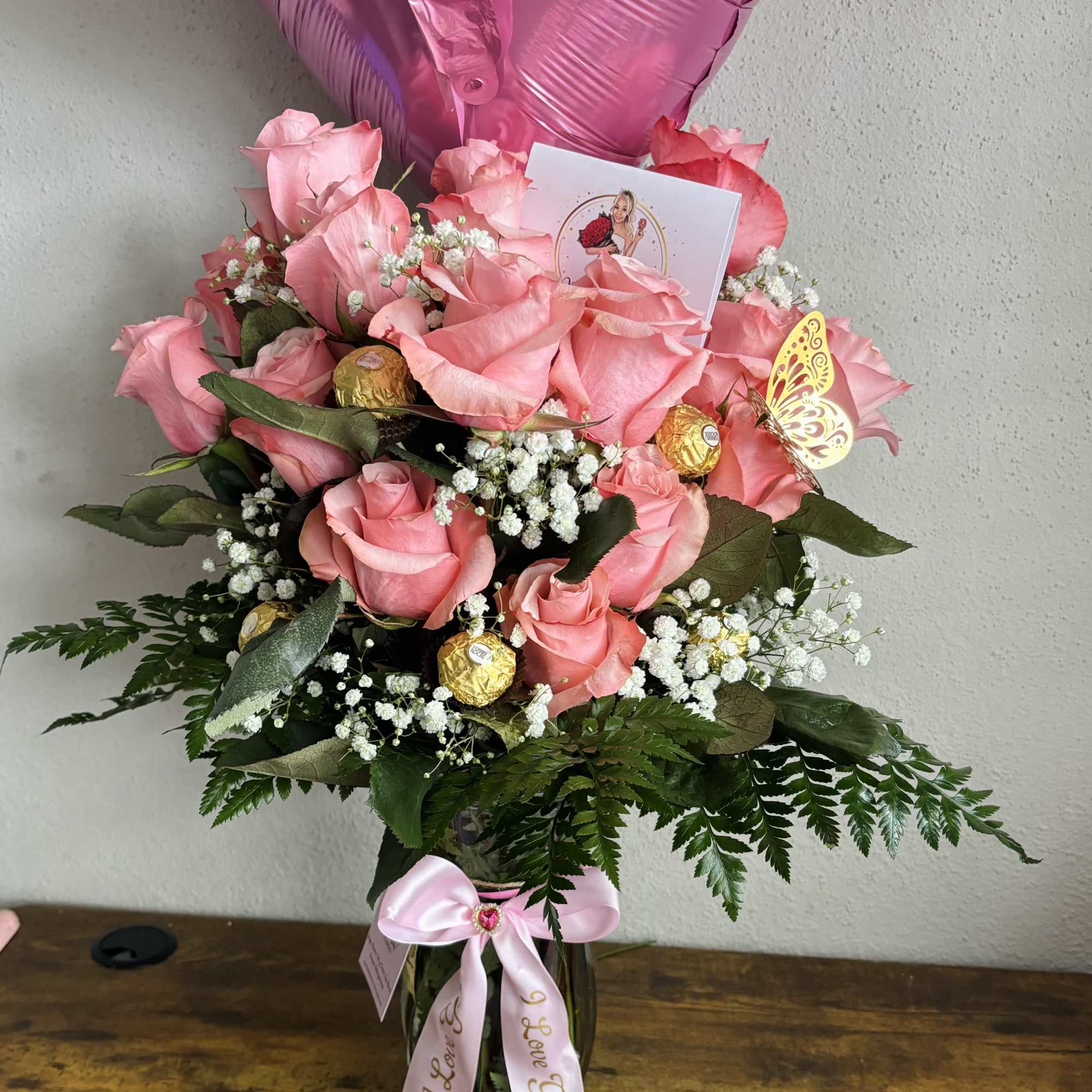 Pink rose bouquet in a glass vase with heart balloons and a ribbon