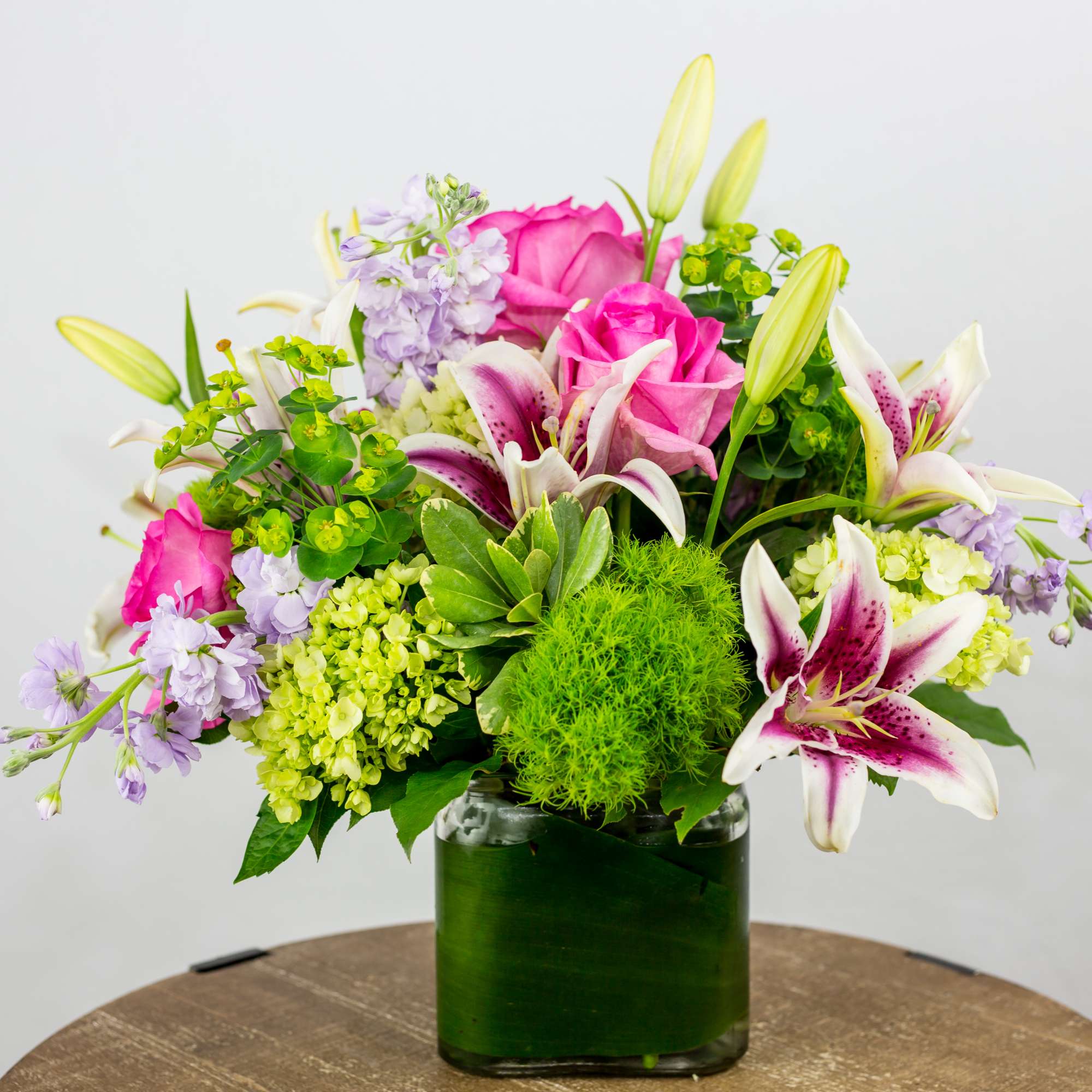 Mixed arrangement with pink lilies, pink roses, and green hydrangea in a leaf-lined glass cube vase