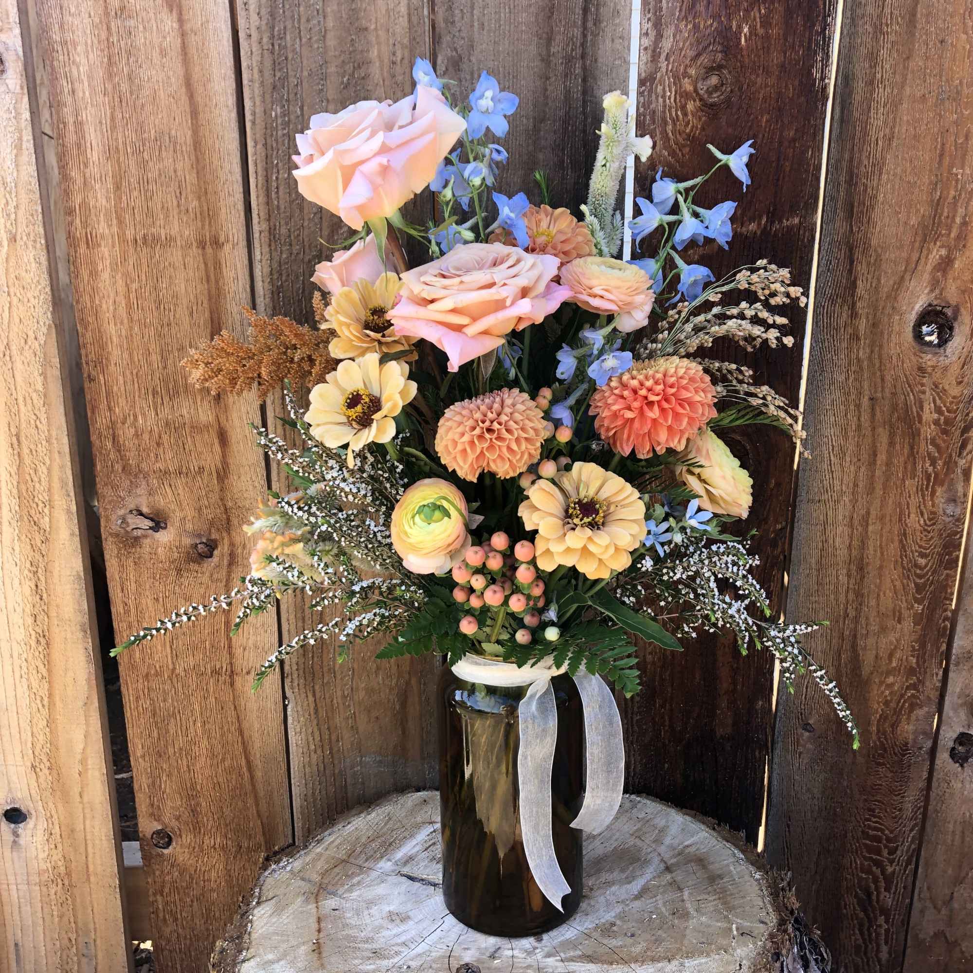 Rustic arrangement of peach roses, dahlias and mixed blooms in an amber glass jar with ribbon.