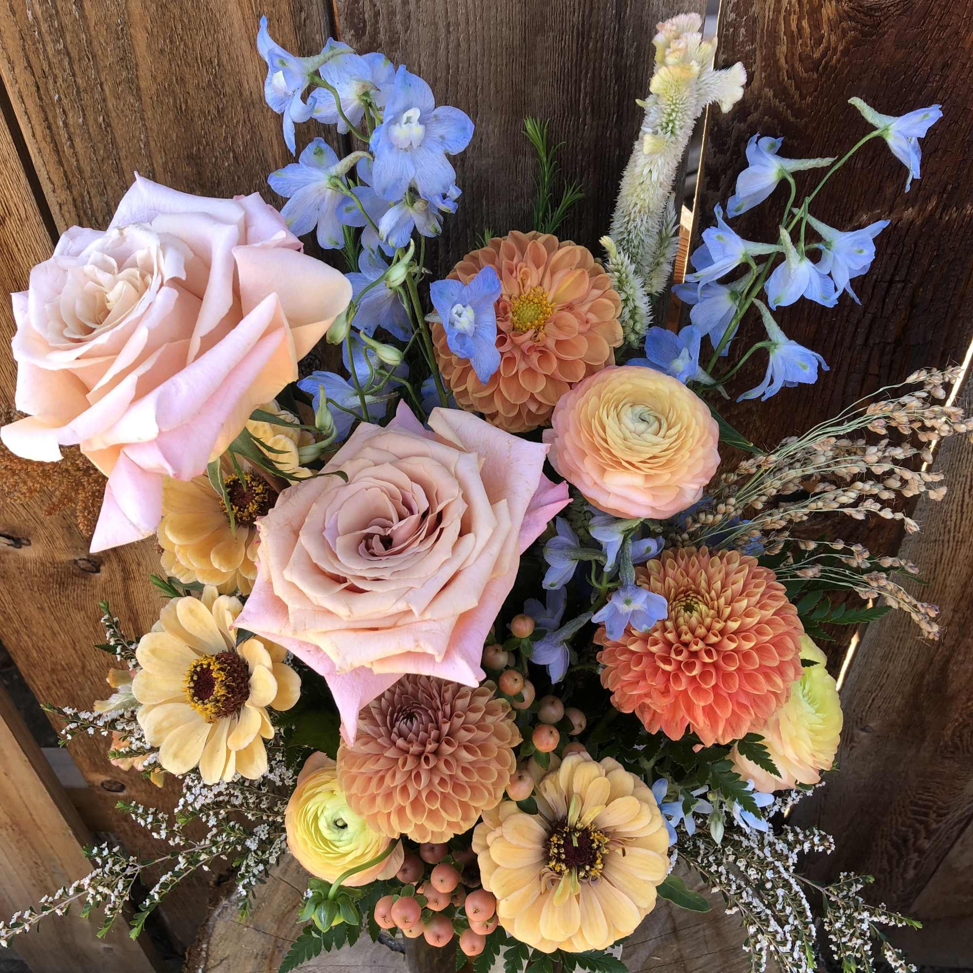 Rustic arrangement of peach roses, orange dahlias, yellow blooms and blue delphinium in a clear vase