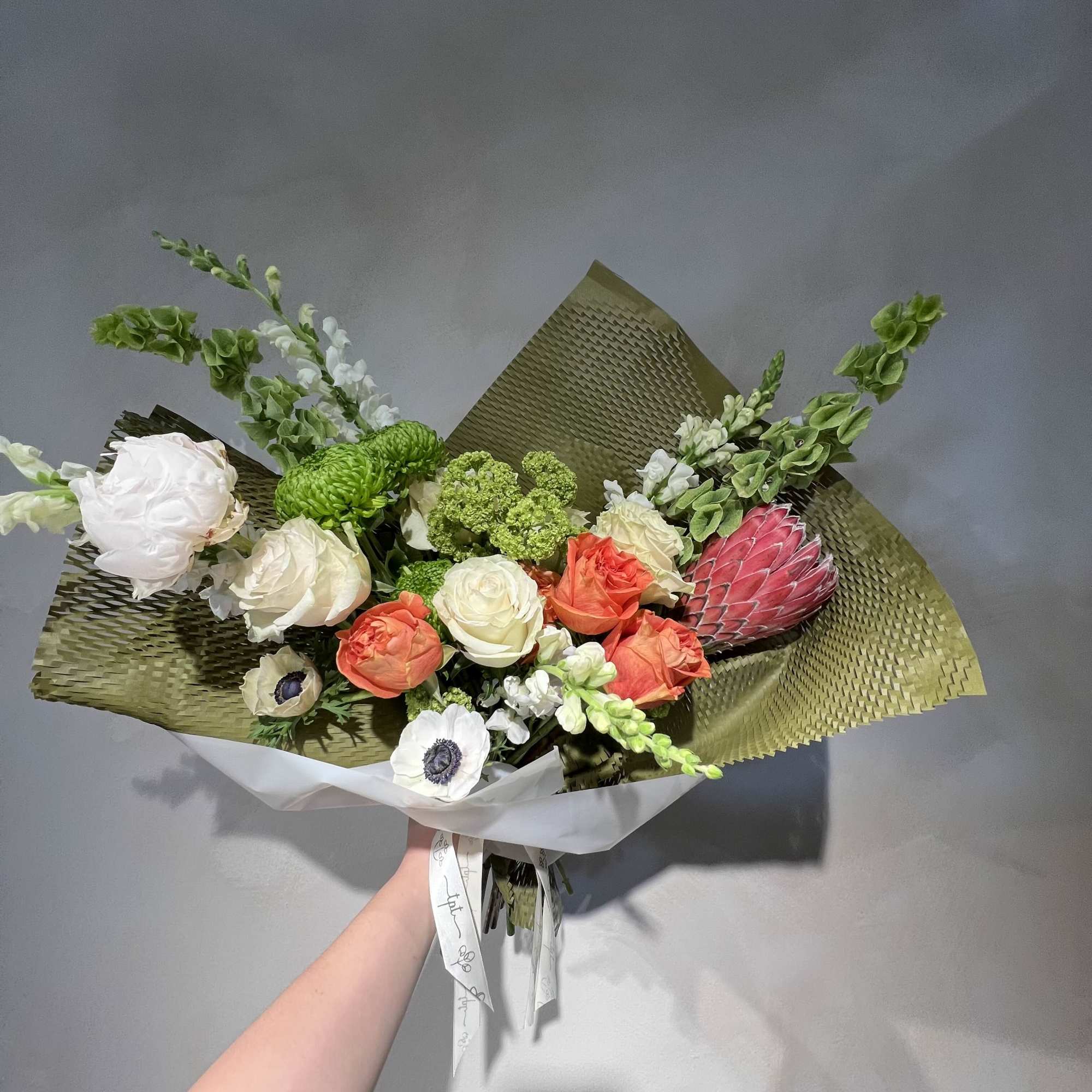 Handheld bouquet of white, coral, and green flowers wrapped in olive paper