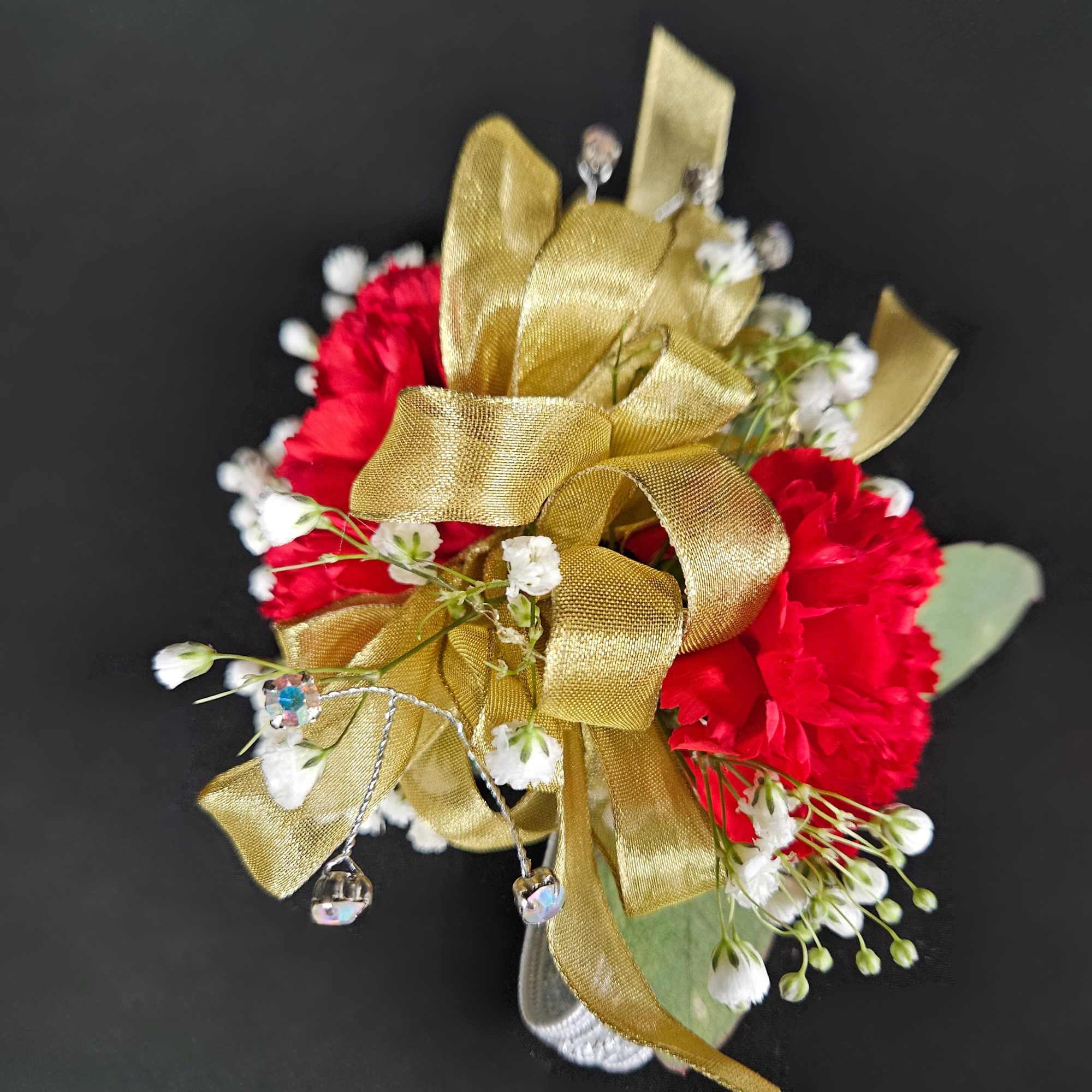 Red carnation corsage with white filler flowers, gold ribbon, and crystal accents on a black background