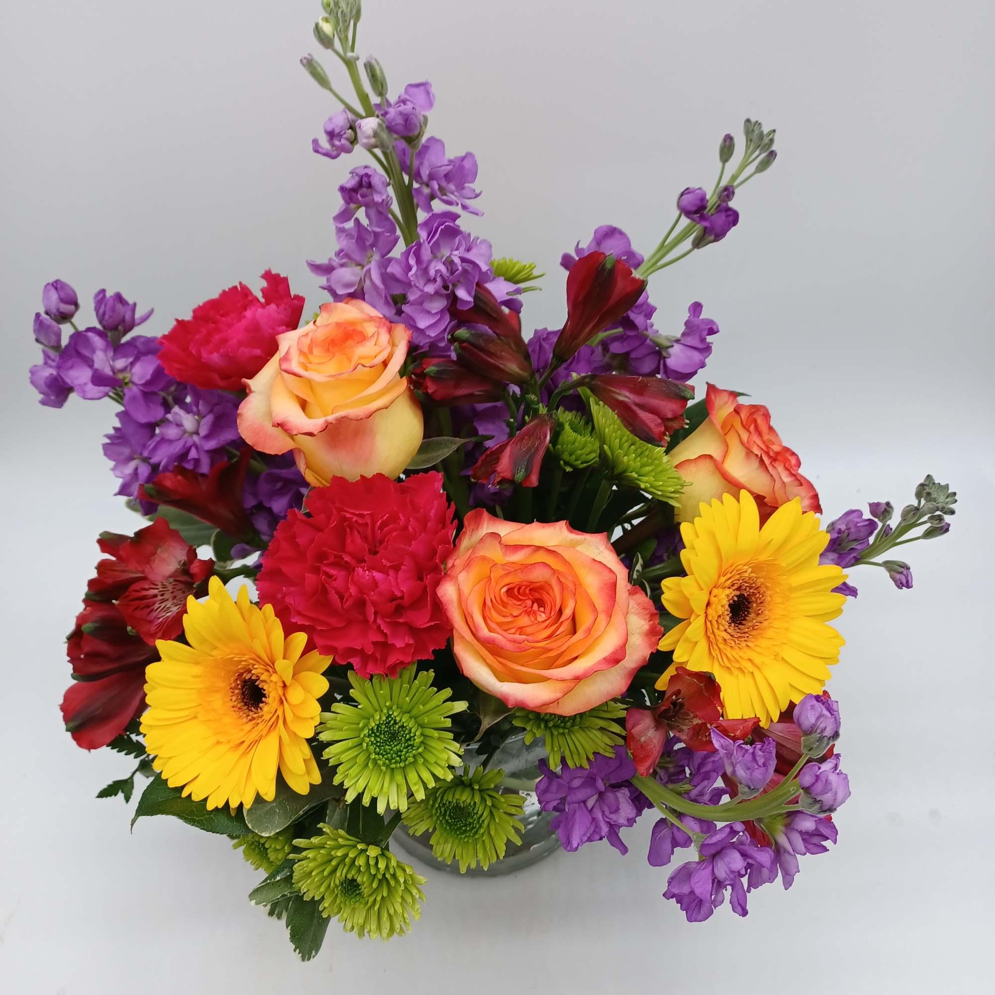 Colorful mixed bouquet with roses, gerbera daisies, carnations, and purple blooms in a glass vase