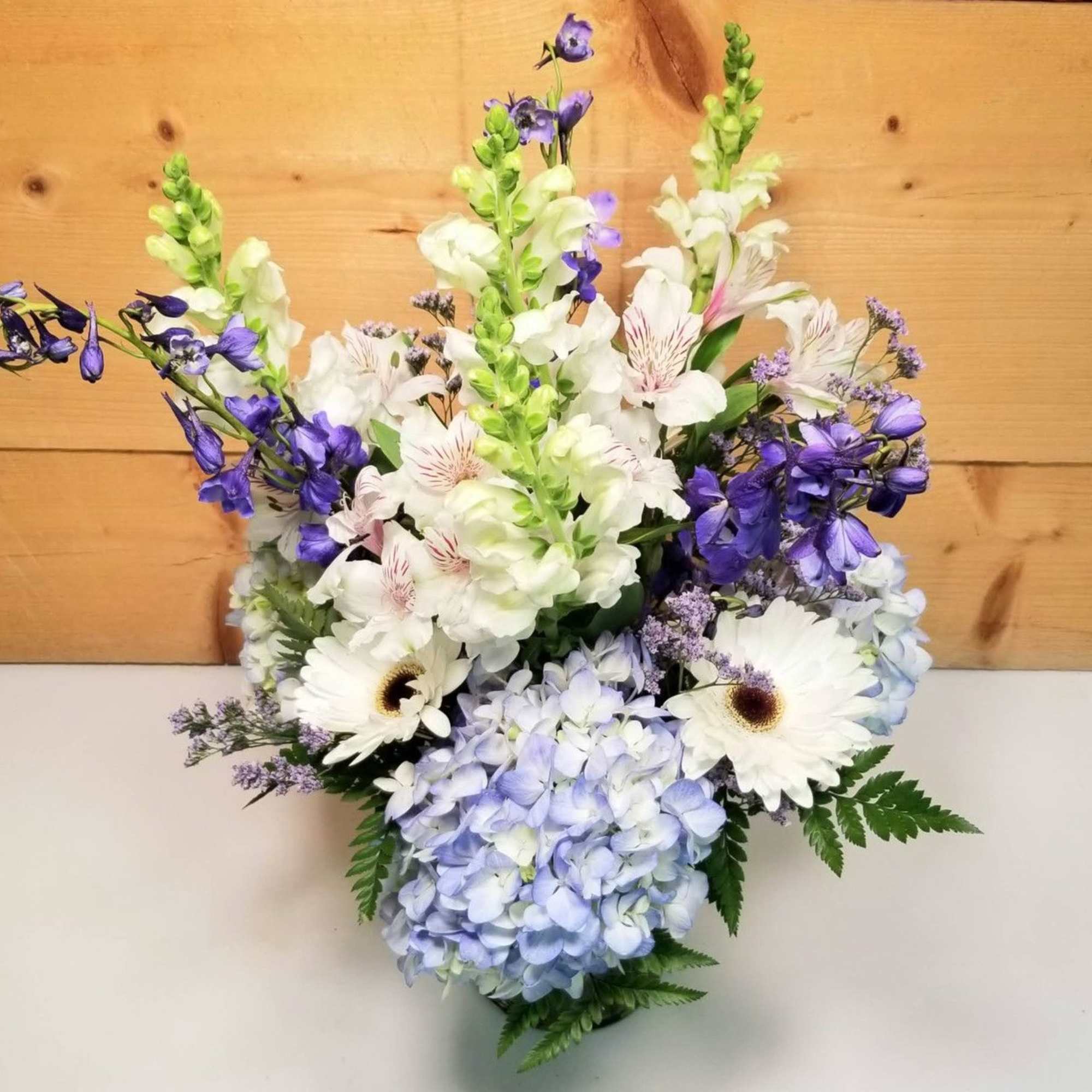 Pastel arrangement of blue hydrangeas, white gerbera daisies, and purple blooms in a clear vase.