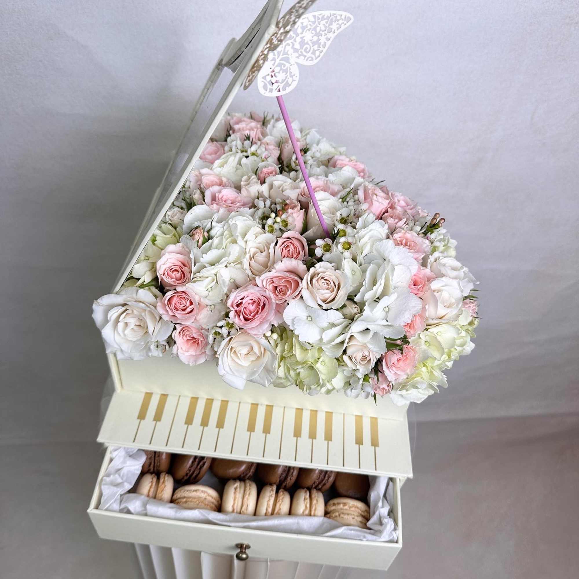 Piano-shaped cream box topped with pink and white roses and hydrangeas, with macarons in a drawer below