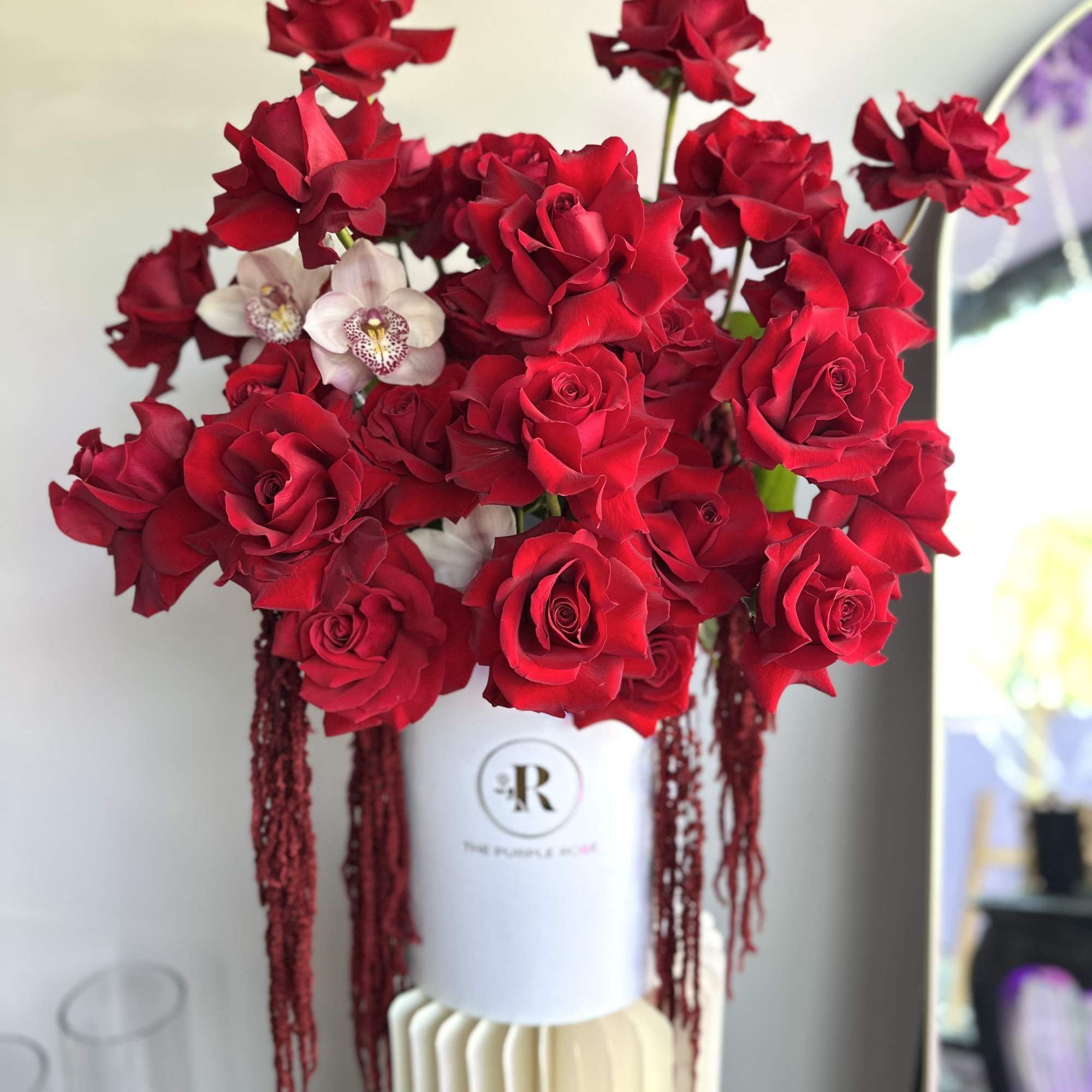 Tall arrangement of red roses with two pale orchids in a white hatbox-style vase on a cream pedestal.